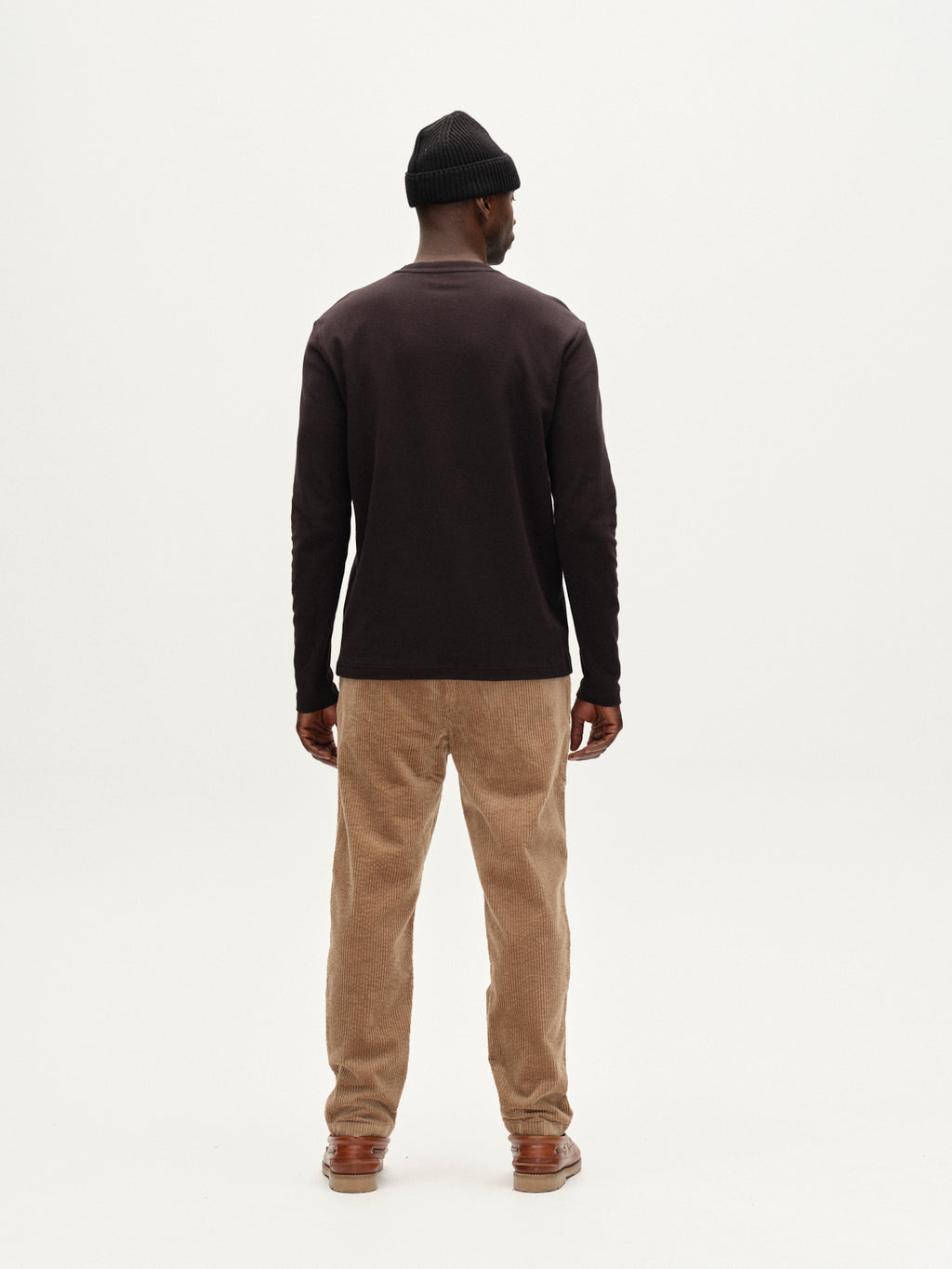 A man stands with his back to the camera in a black beanie, long-sleeve shirt, brown shoes, and Gabba's Monza Ibre - Sand beige corduroy comfort stretch trousers against a plain white background.