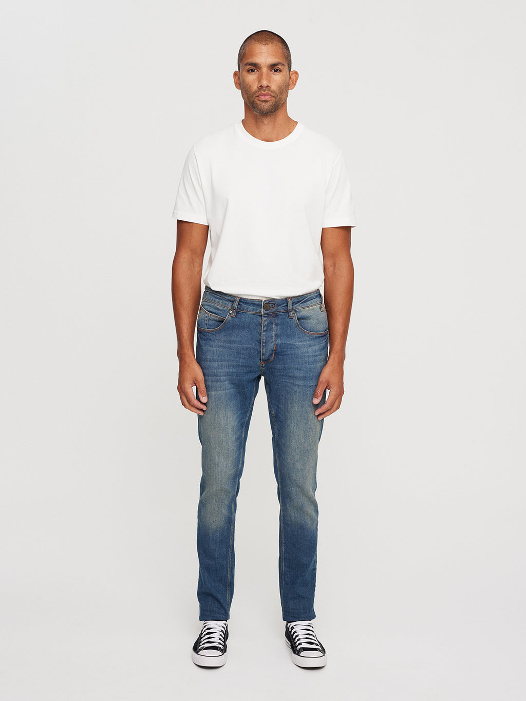 A man stands against a plain background in a relaxed look with a white T-shirt, GABBAs Rey 44617 Jeans in denim wash, and black sneakers with white soles. He has short hair and a neutral expression.