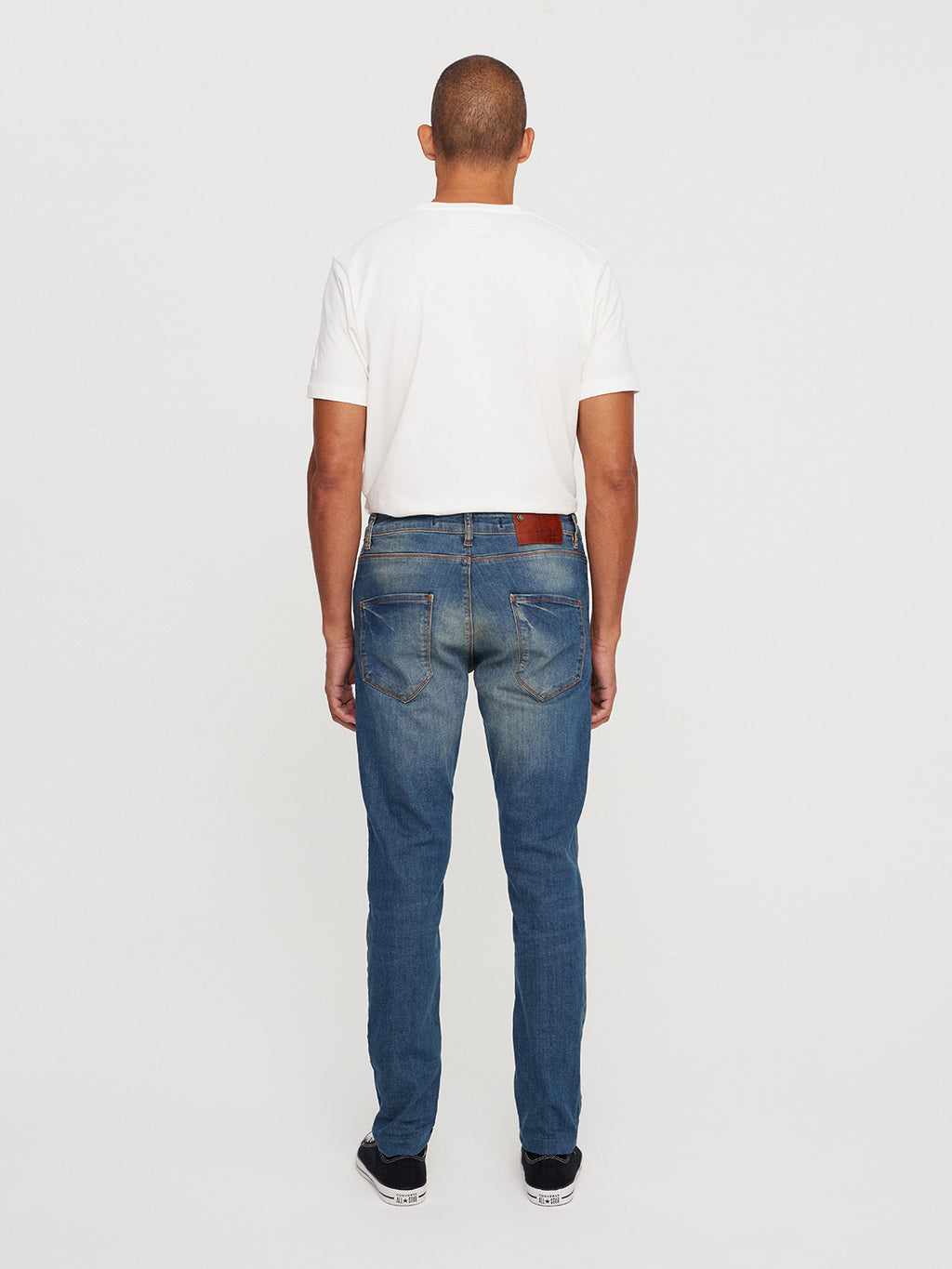 A man wearing a white t-shirt, GABBAs Rey 44617 Jeans in denim wash, and black shoes stands with his back to the camera against a plain white background.
