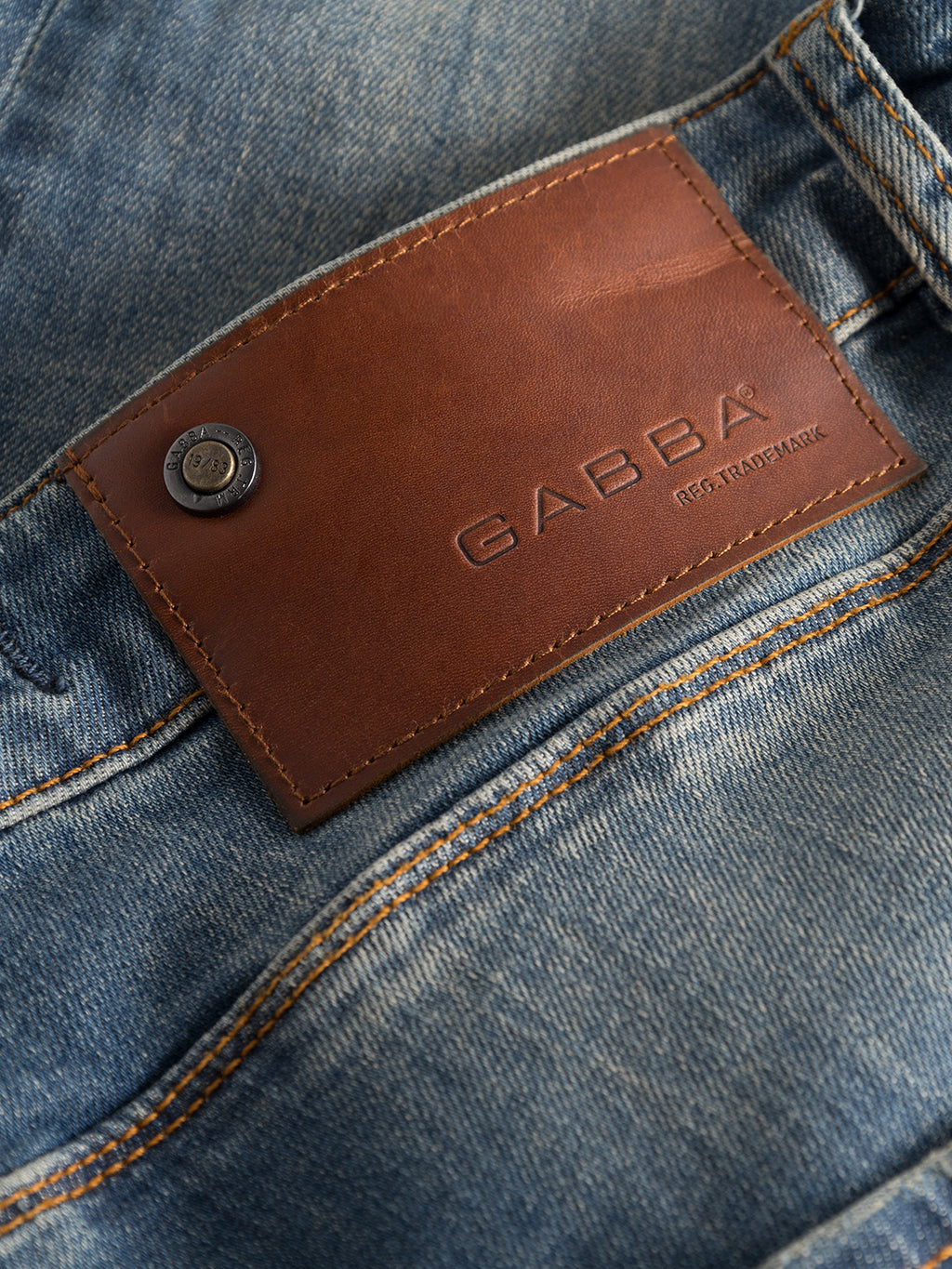 Close-up of Rey 44617 Jeans - Denim wash by GABBA showcases a brown leather patch labeled REG. TRADEMARK, with an adjacent metal button, enhancing the slim jeans casual vibe. Orange stitching along the seams completes the look.