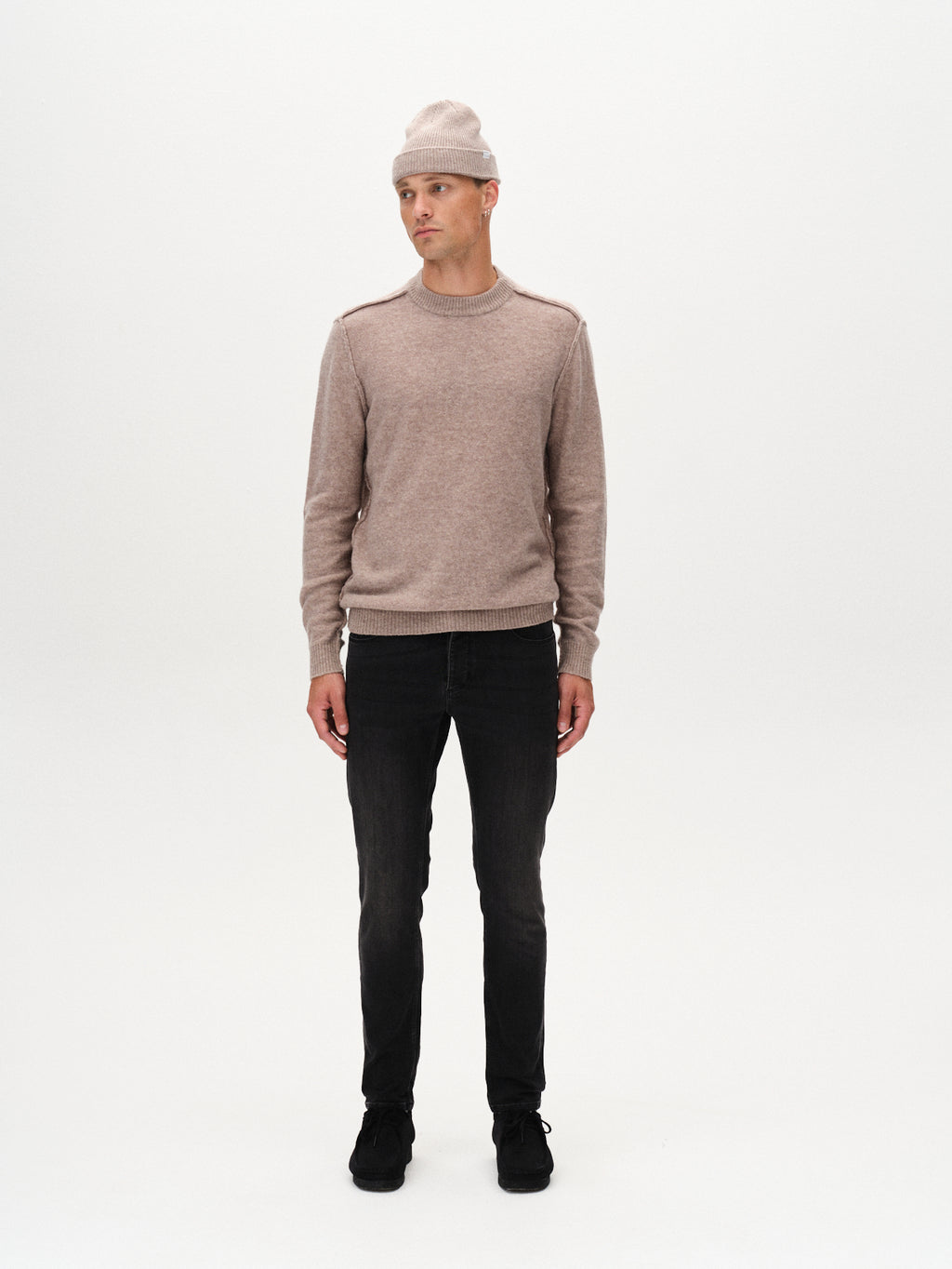 A man stands facing forward against a white background, wearing a beige knit beanie, matching sweater, GABBA Rey Thor Jeans in Denim wash (slim fit), and black shoes for a laid-back look.