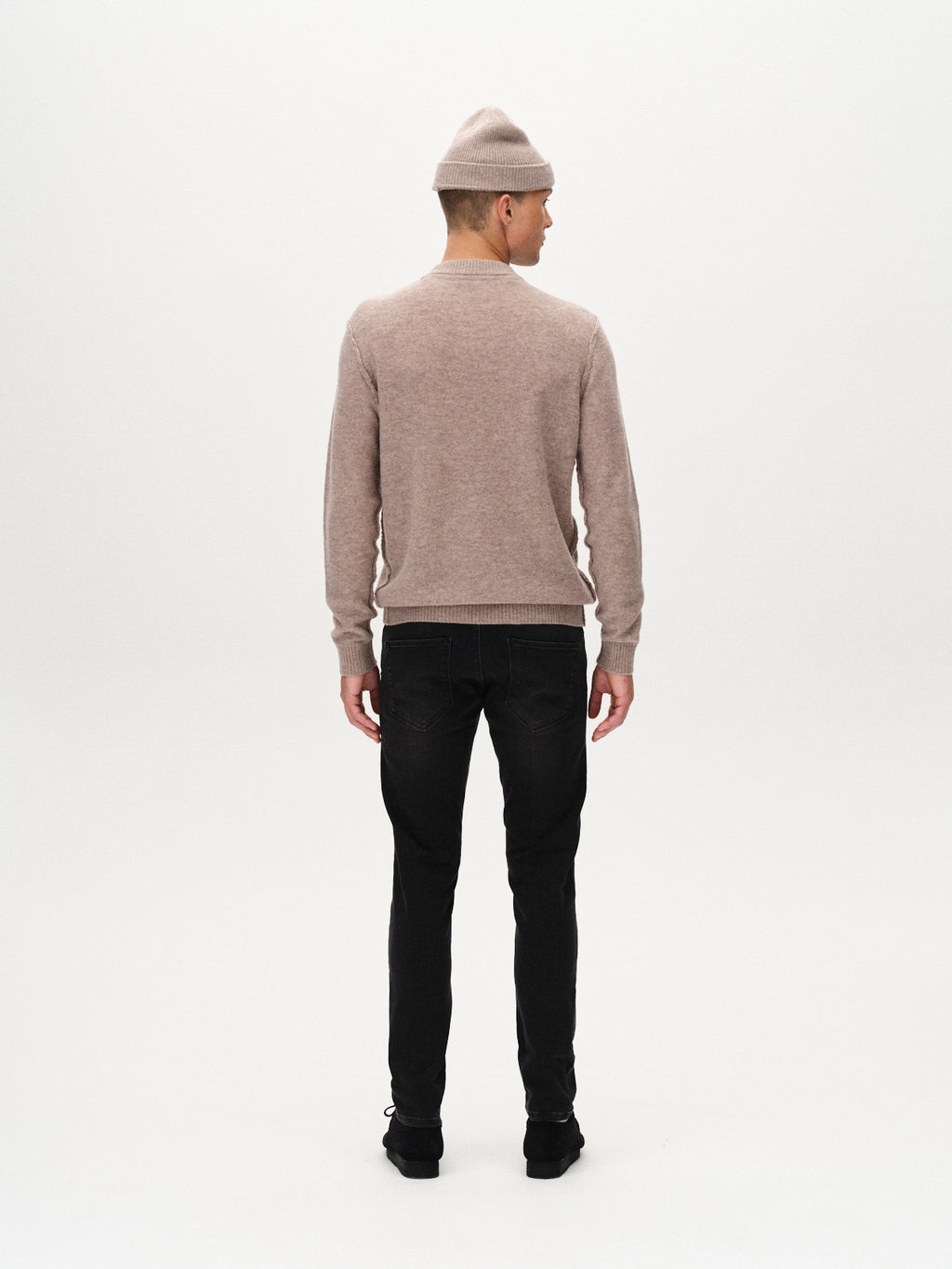 A person stands facing away against a plain white background, wearing a beige beanie, light brown sweater, and GABBA Rey Thor Jeans - Denim wash with black shoes for a laid-back look.