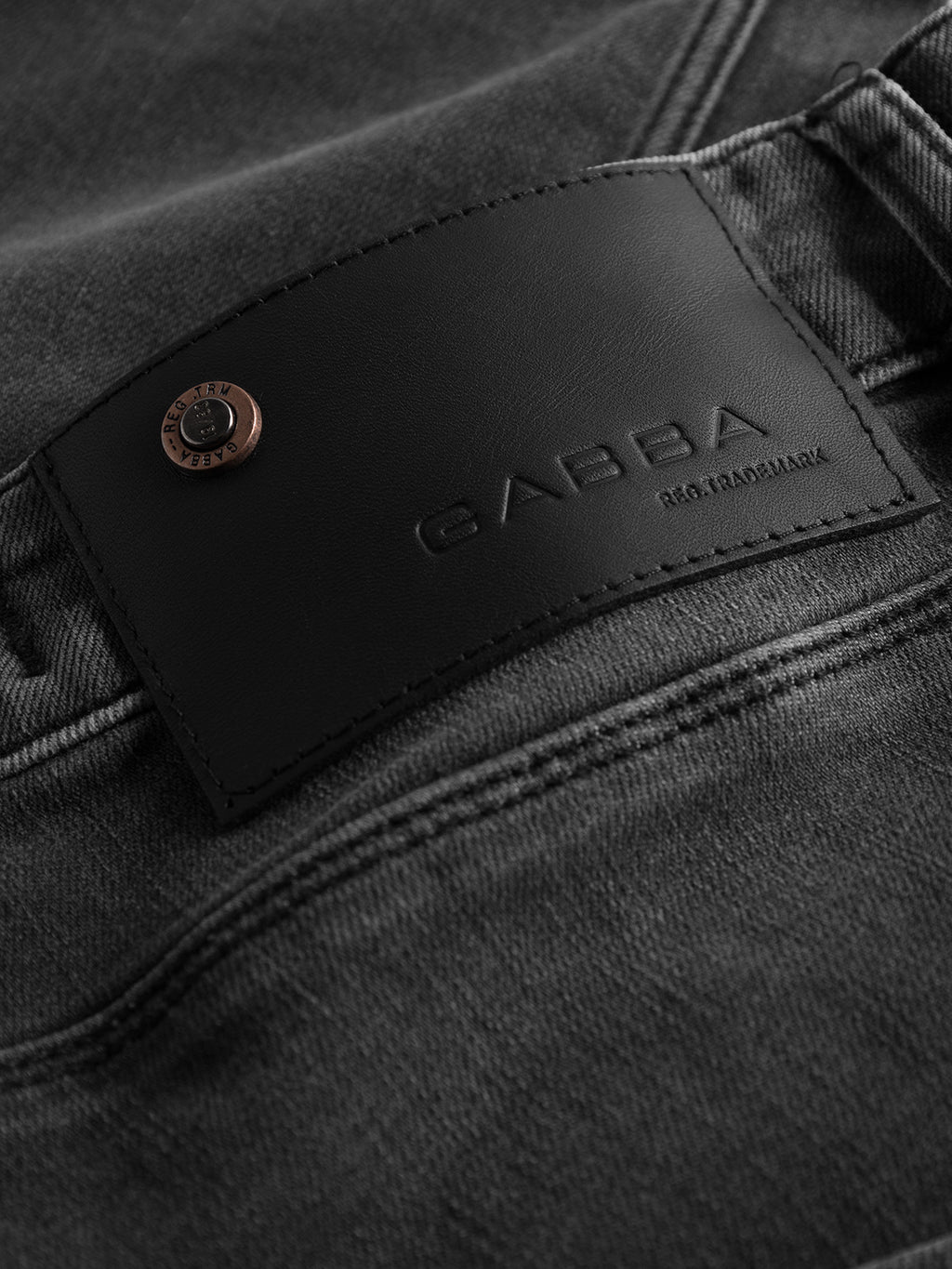 Close-up of dark gray denim fabric from the Rey Thor Jeans - Denim wash collection, featuring a leather patch embossed with the brand name GABBA. A bronze rivet adds a touch of flair, making these jeans perfect for achieving a laid-back slim fit style.
