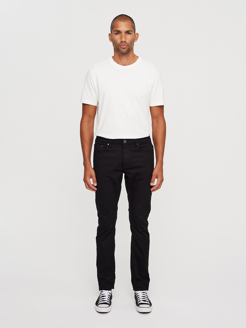 Facing the camera against a plain background, a person wears a white T-shirt and GABBAs Jones K1911 Black Jeans in black denim, paired with black and white sneakers. Their arms rest at their sides with a neutral expression.