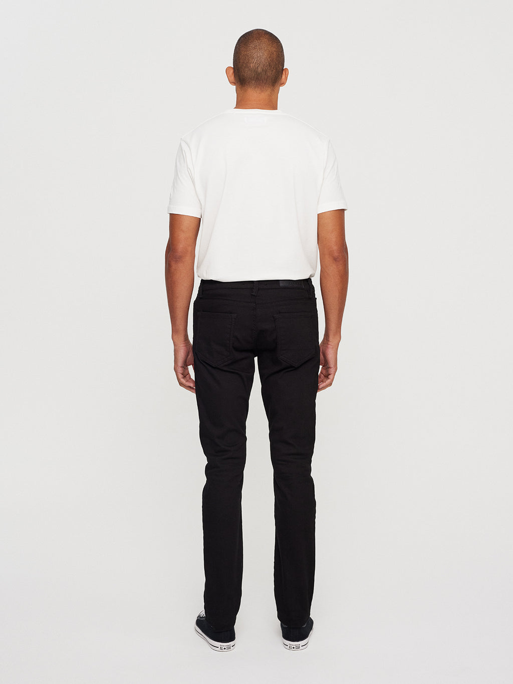 A person with a slim silhouette wears a white T-shirt, GABBAs Jones K1911 Black Jeans in black denim, and black sneakers with white soles against a plain, light background.