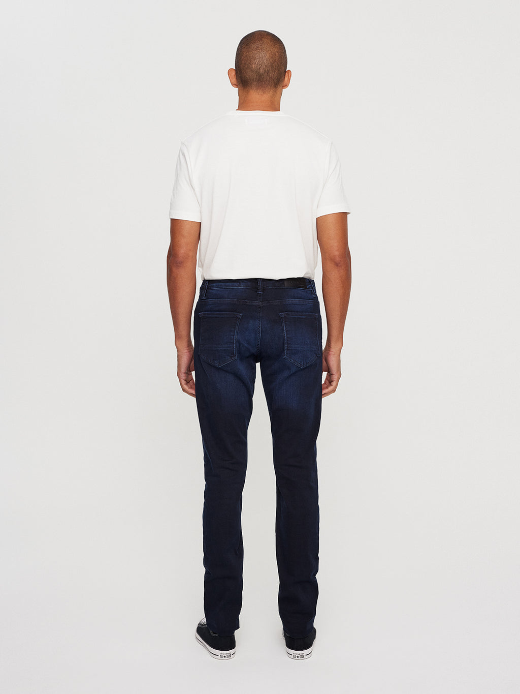 Standing with their back to the camera, a person wears a white T-shirt, GABBAs Jones K2291 - Denim wash slim silhouette jeans made from super-stretch fabric, and black shoes against a plain light background.