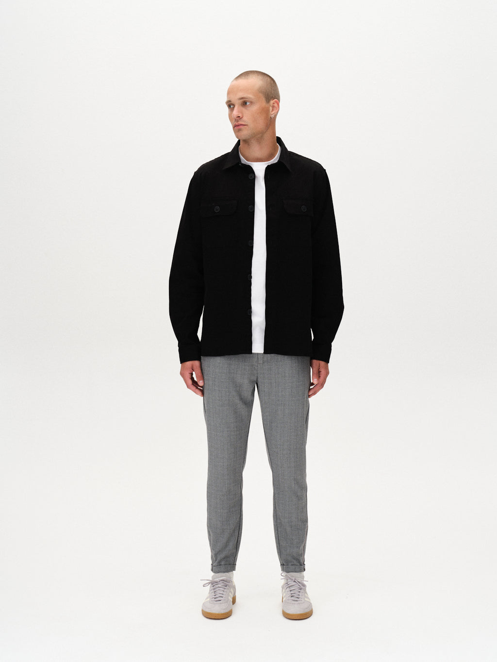 A man stands against a white background wearing the GABBA Pisa Cross Lt Grey pants, a black jacket over a white shirt, and white sneakers. He looks slightly to his left with his arms relaxed at his sides.