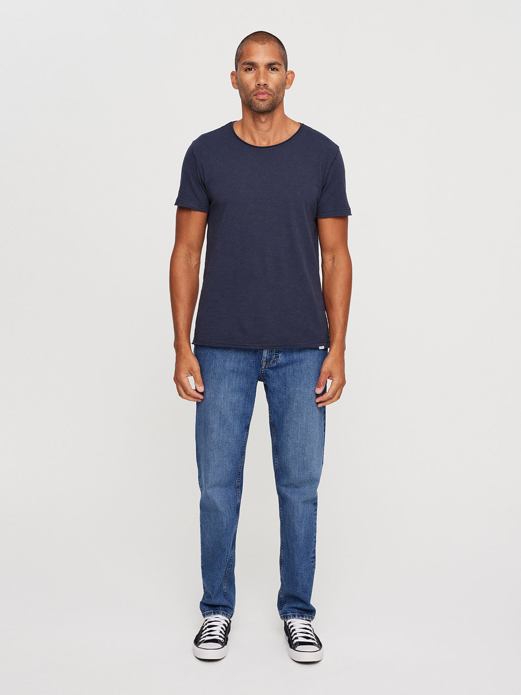 A man wearing the GABBA Konrad Slub S/S Tee in navy, comfortable blue jeans, and black sneakers with white soles stands against a plain white background. His neutral expression and relaxed arms effortlessly highlight the tees regular fit.