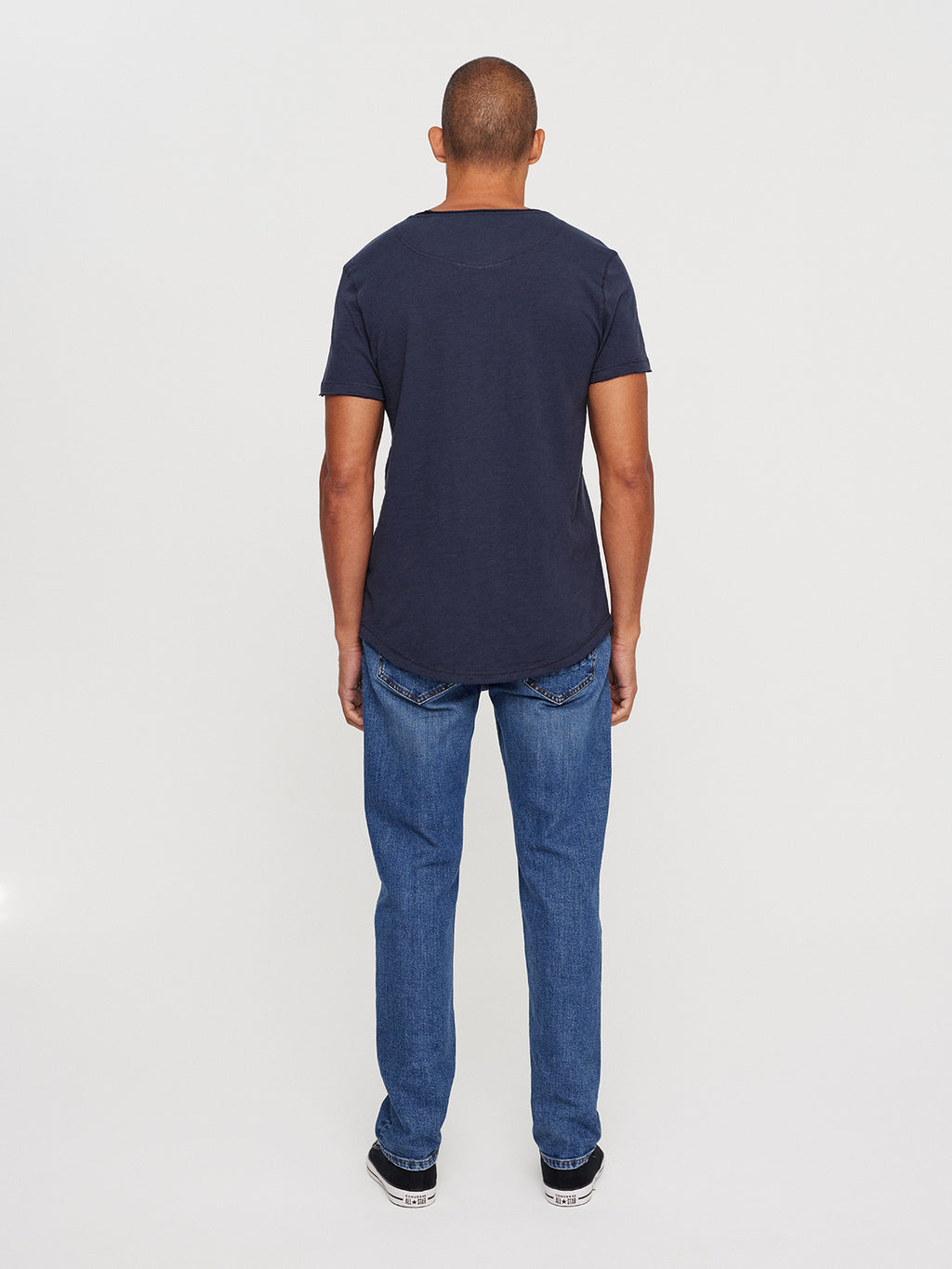 A person stands facing away, wearing the GABBA Konrad Slub S/S Tee in navy with light blue jeans and black sneakers. The flawless white background accentuates the outfits casual charm.