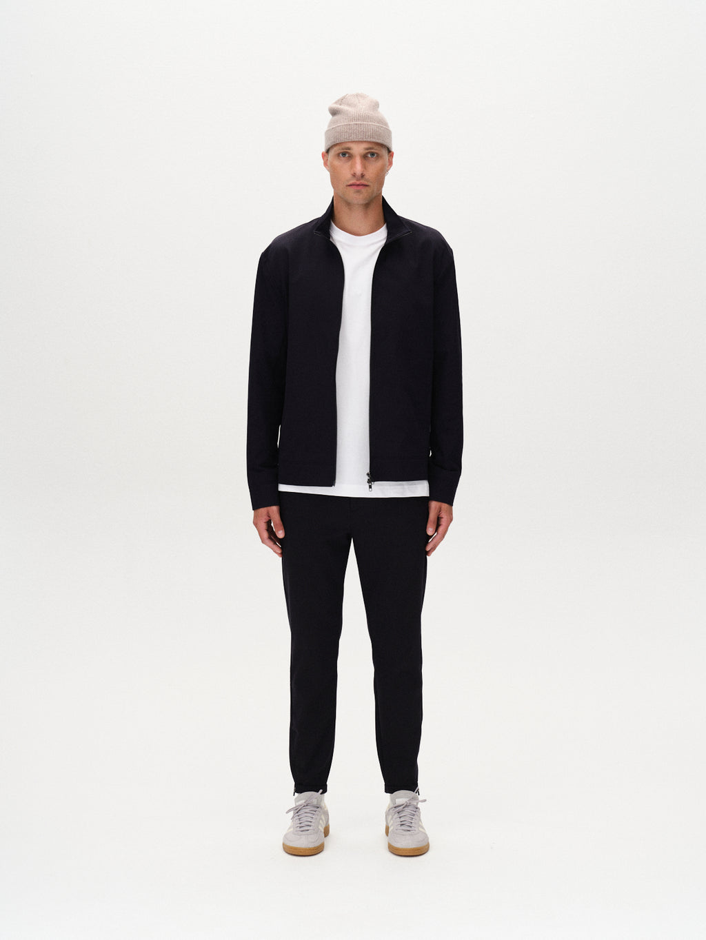 A man faces forward against a white background, wearing the GABBA Pisa Small Dot - Navy zip-up jacket, a beige beanie, white T-shirt, black metro-style pants, and white sneakers. His arms are relaxed at his sides.