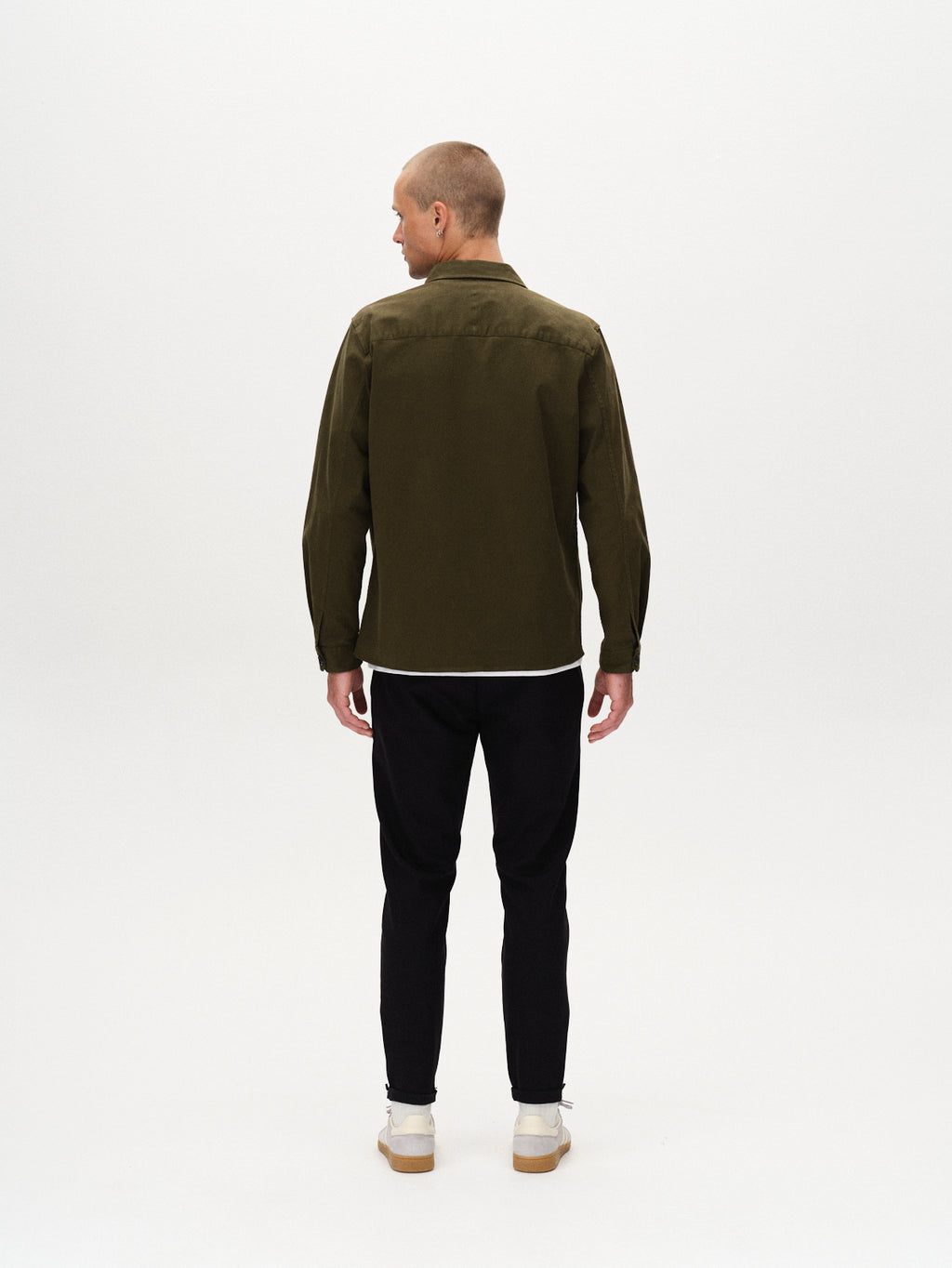 A man with a shaved head stands facing away, wearing black GABBA Pisa Small Dot pants, an olive green jacket, and white sneakers against a plain white background.