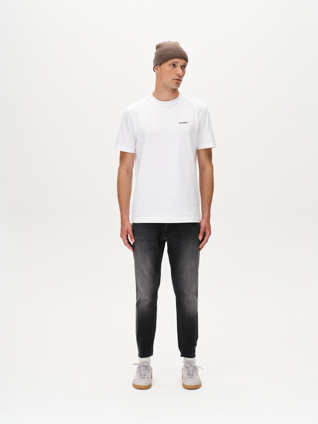 A man stands facing forward against a white background, wearing a gray beanie, plain white T-shirt, gray sneakers, and GABBA Alex Thor Cropped Jeans in denim wash.