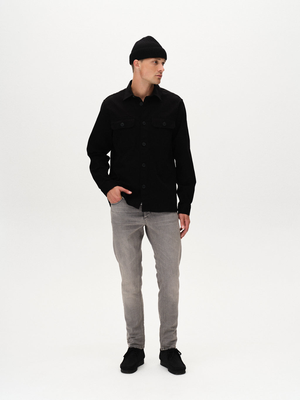 A man stands against a plain white background, looking to his left with one hand in his pocket. He wears a black beanie, black button-up shirt, black shoes, and slim fit GABBA Rey K3454 - Denim wash jeans for a laid-back look.