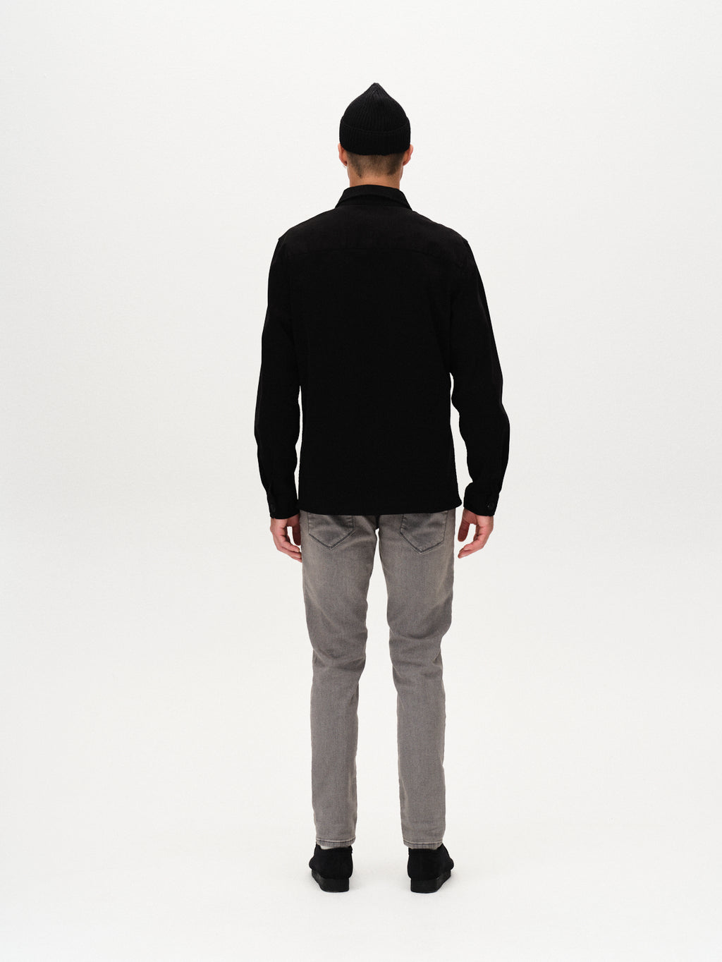 A person stands facing away from the camera against a white background, wearing a black long-sleeve shirt, GABBA Rey K3454 - Denim wash slim fit gray jeans, black shoes, and a black beanie for a relaxed vibe.
