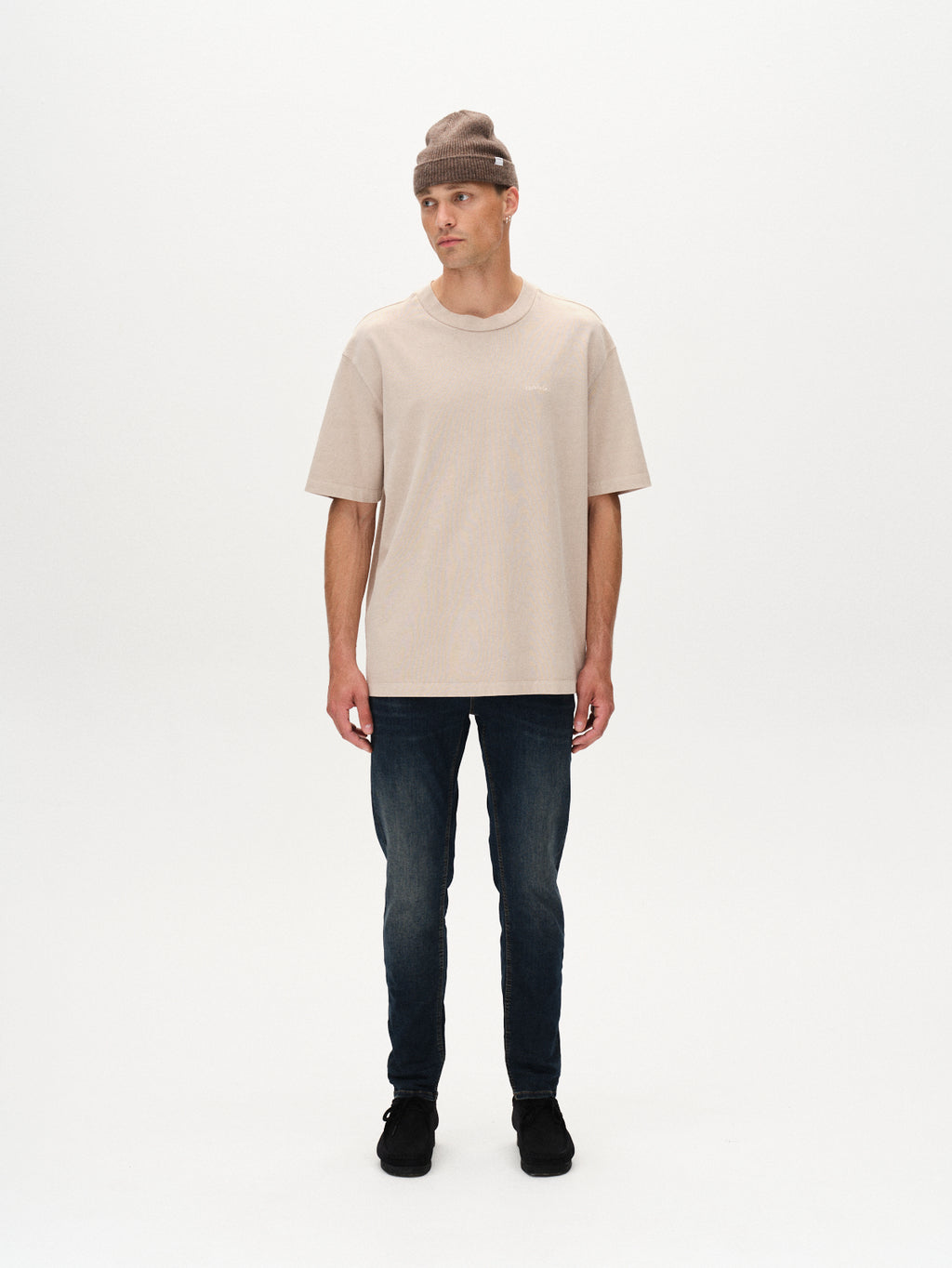 A man in a beige beanie, beige oversized tee, GABBA Rey K3606 Mid Blue Jeans in mid blue denim, and black shoes stands against a plain white background, looking slightly to his left for a laid-back vibe.
