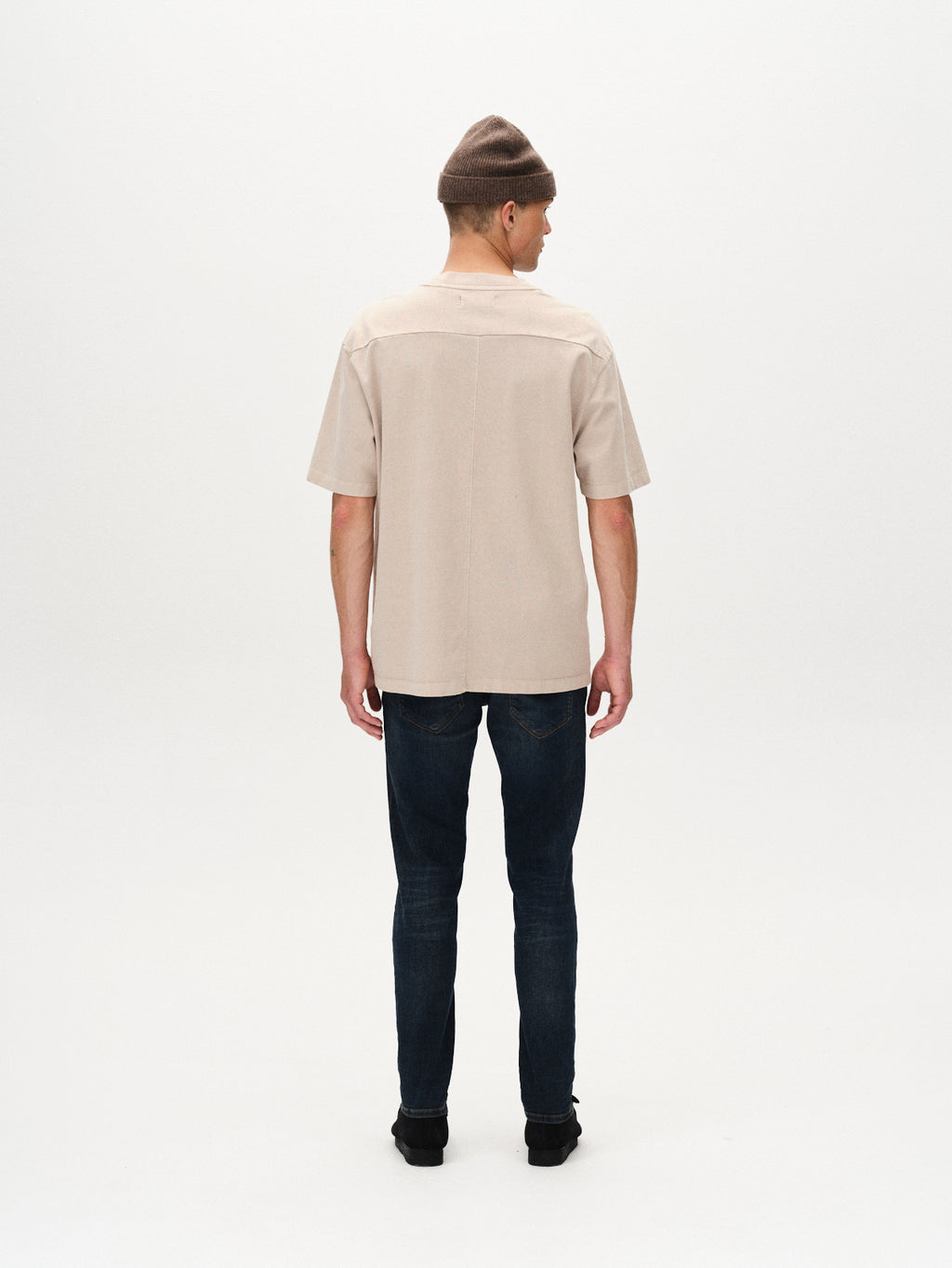 A person stands facing away from the camera in a brown beanie, beige short-sleeve shirt, GABBA Rey K3606 Mid Blue Jeans in mid blue denim, and black shoes against a plain white background for a laid-back look.