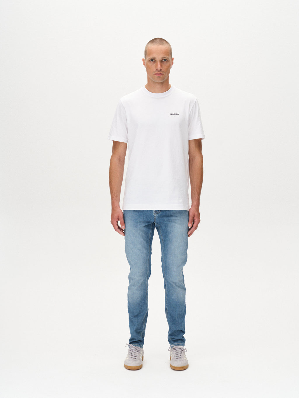 A person faces forward against a plain white background, wearing GABBAs Jones K2615 Lt. - Denim wash jeans with a slim fit, styled with a white t-shirt and gray sneakers. Their arms are relaxed by their sides and their expression is neutral.