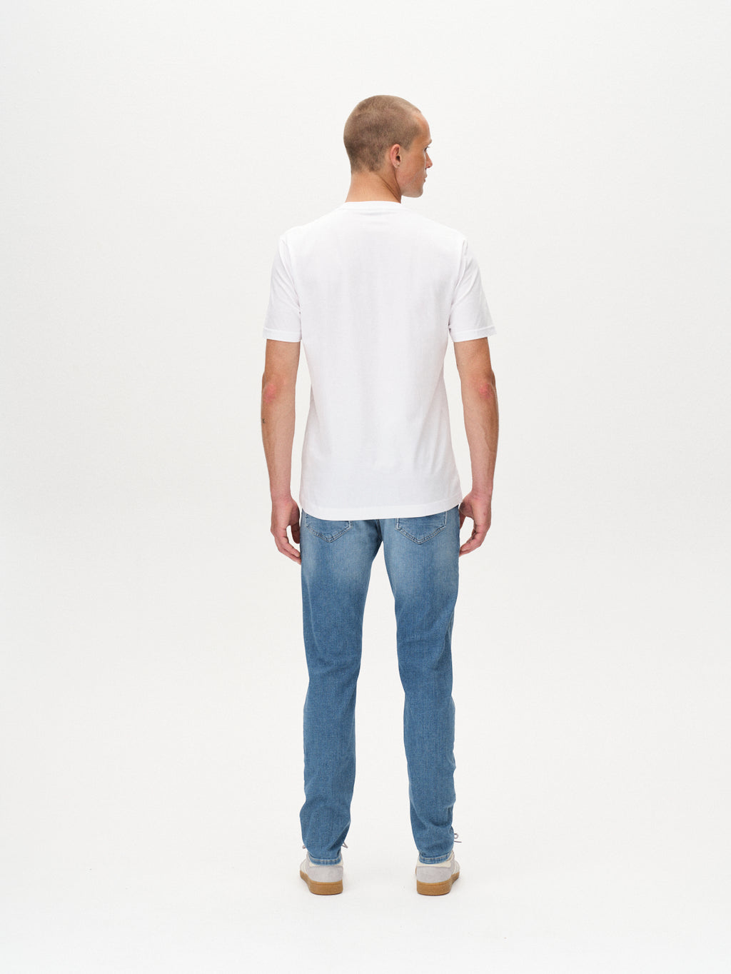 A man faces away from the camera, wearing a plain white t-shirt, GABBA Jones K2615 Lt. - Denim wash slim light blue jeans, and white sneakers against a white background.