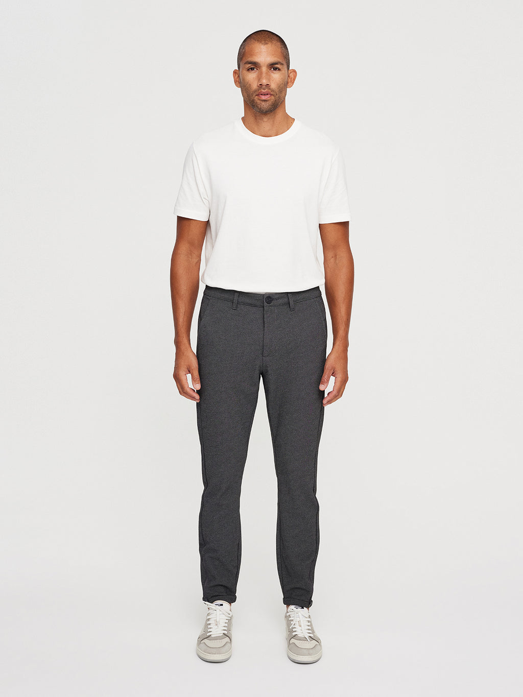 A man stands against a plain background in a white T-shirt and GABBAs Pisa Jersey Pant in Grey Mel, paired with light gray sneakers, exuding a clean metro look. He has close-cropped hair with his hands at his sides.