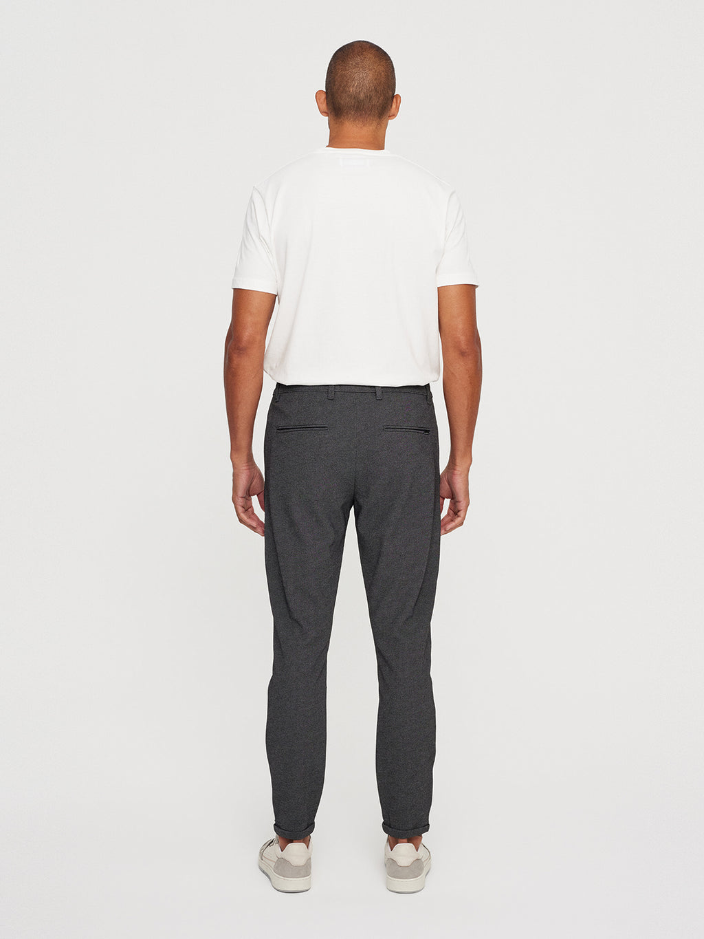 A person with a metro look is facing away, wearing a white t-shirt paired with GABBAs Pisa Jersey Pant in Grey Mel and white sneakers, exuding a modern vibe against the plain, light-colored background.
