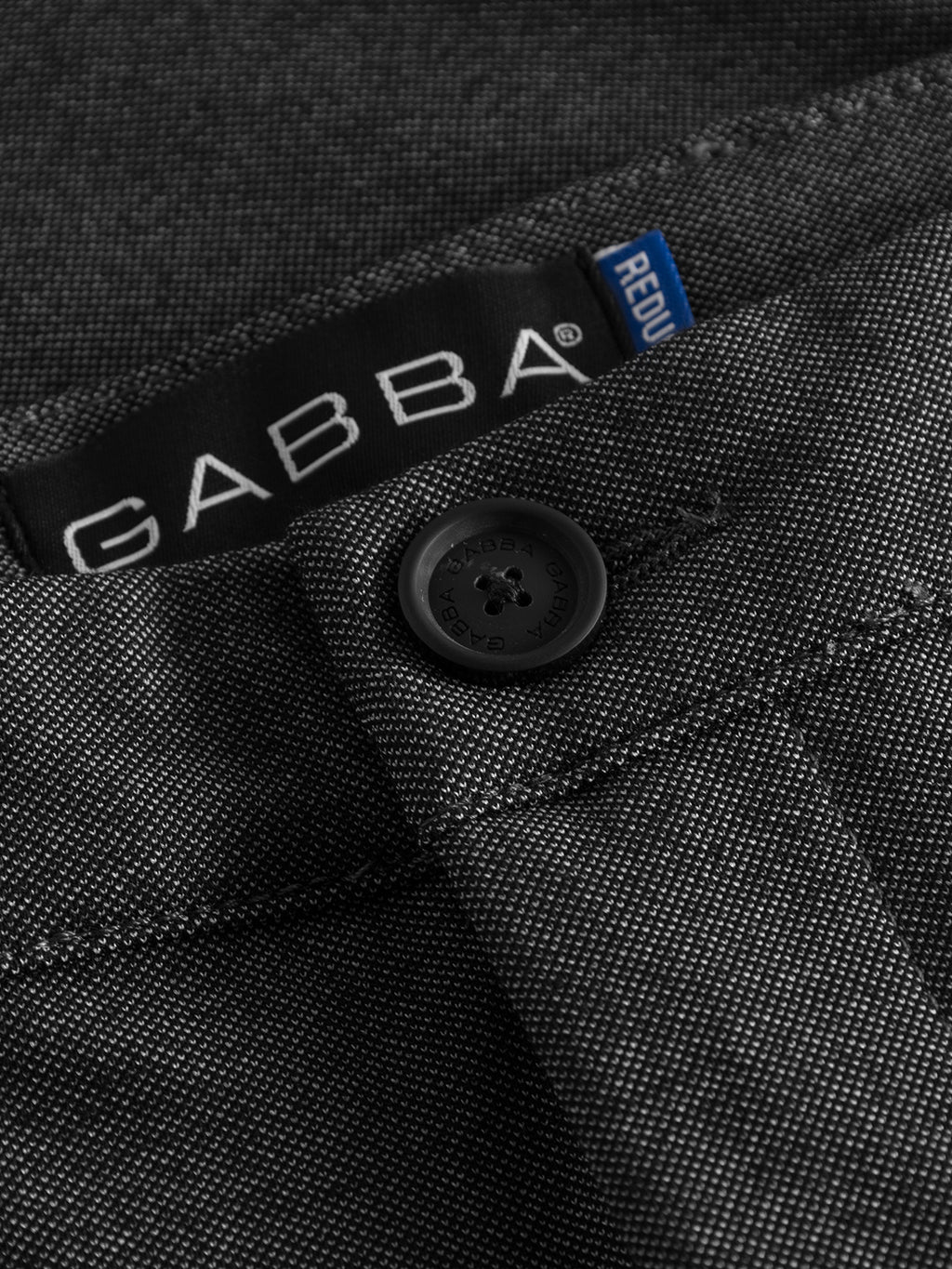 Close-up of the Pisa Jersey Pant in Grey Mel by GABBA, featuring a tapered fit with a visible button and stitching. The brand label is partially visible above the waistband with a blue tag. Made from high-stretch fabric, the textured material enhances its metro look.