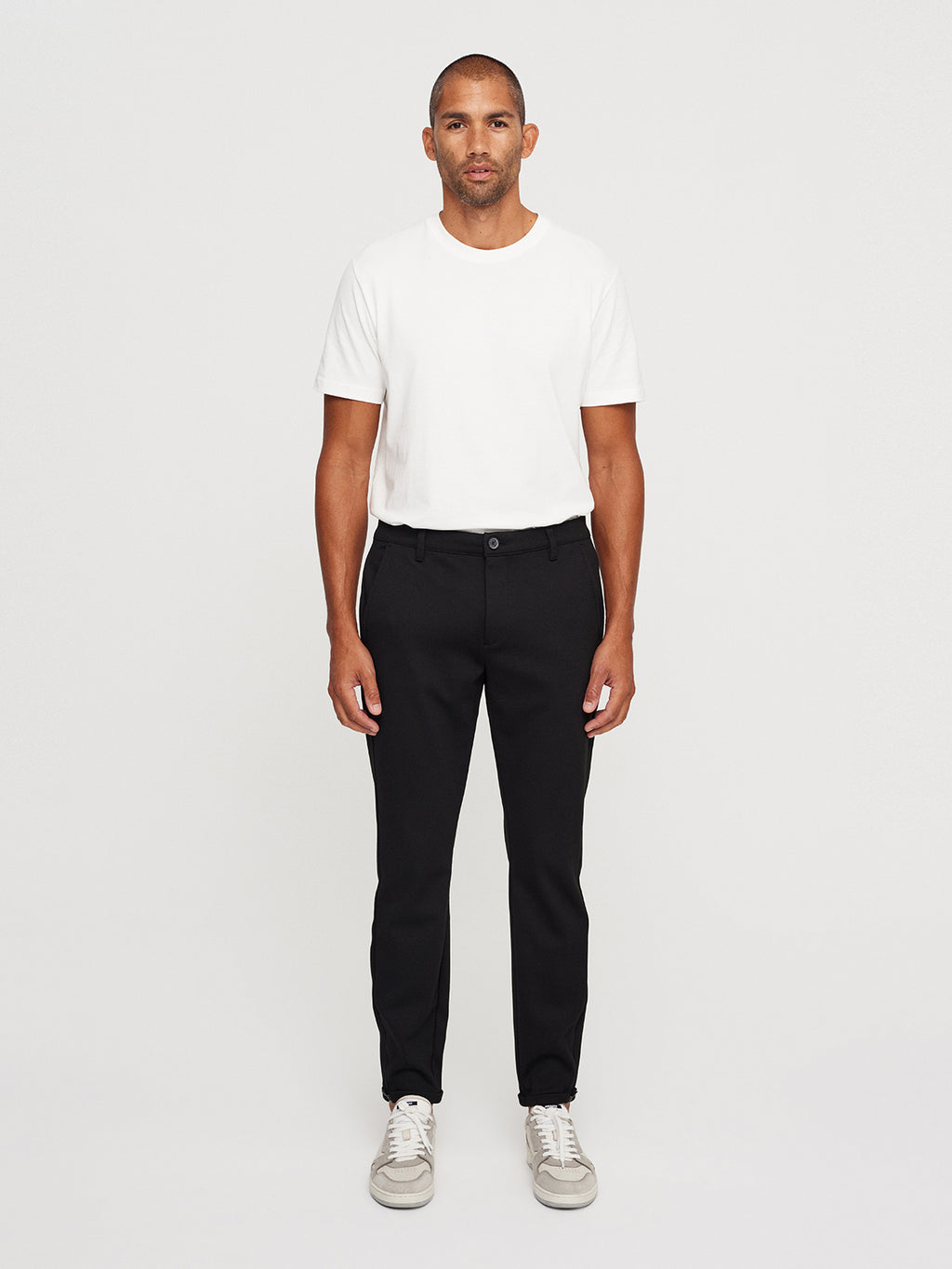 A man with short hair stands against a plain background in a white T-shirt, Pisa Jersey Pant - Black by GABBA, and white sneakers, looking directly at the camera with a neutral expression, embodying effortless metro style.