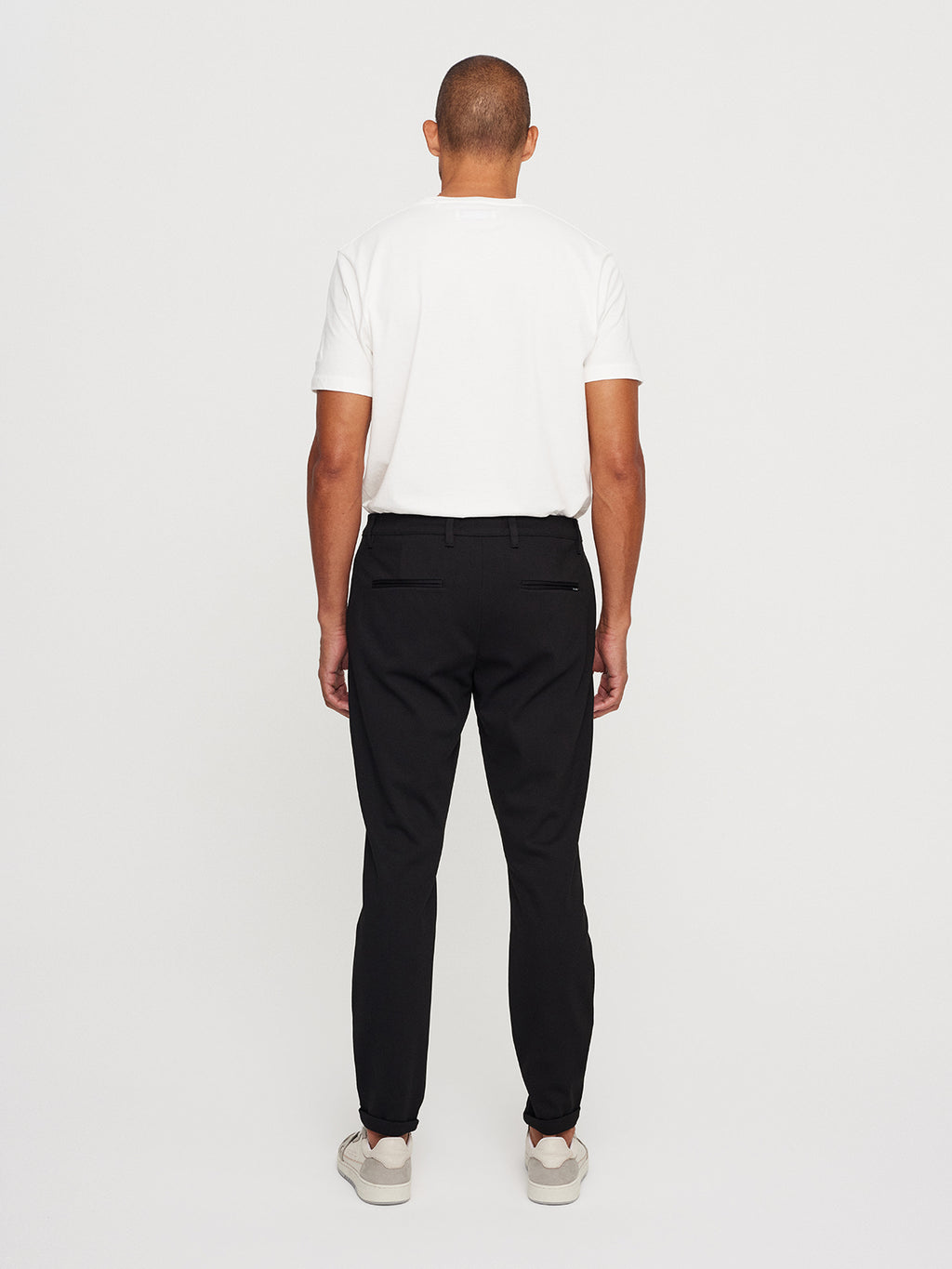A person stands facing away, showcasing a sleek metro vibe with a white T-shirt, Pisa Jersey Pant in black by GABBA for a tapered fit, and white sneakers. The plain white background enhances the casual yet stylish ensemble.