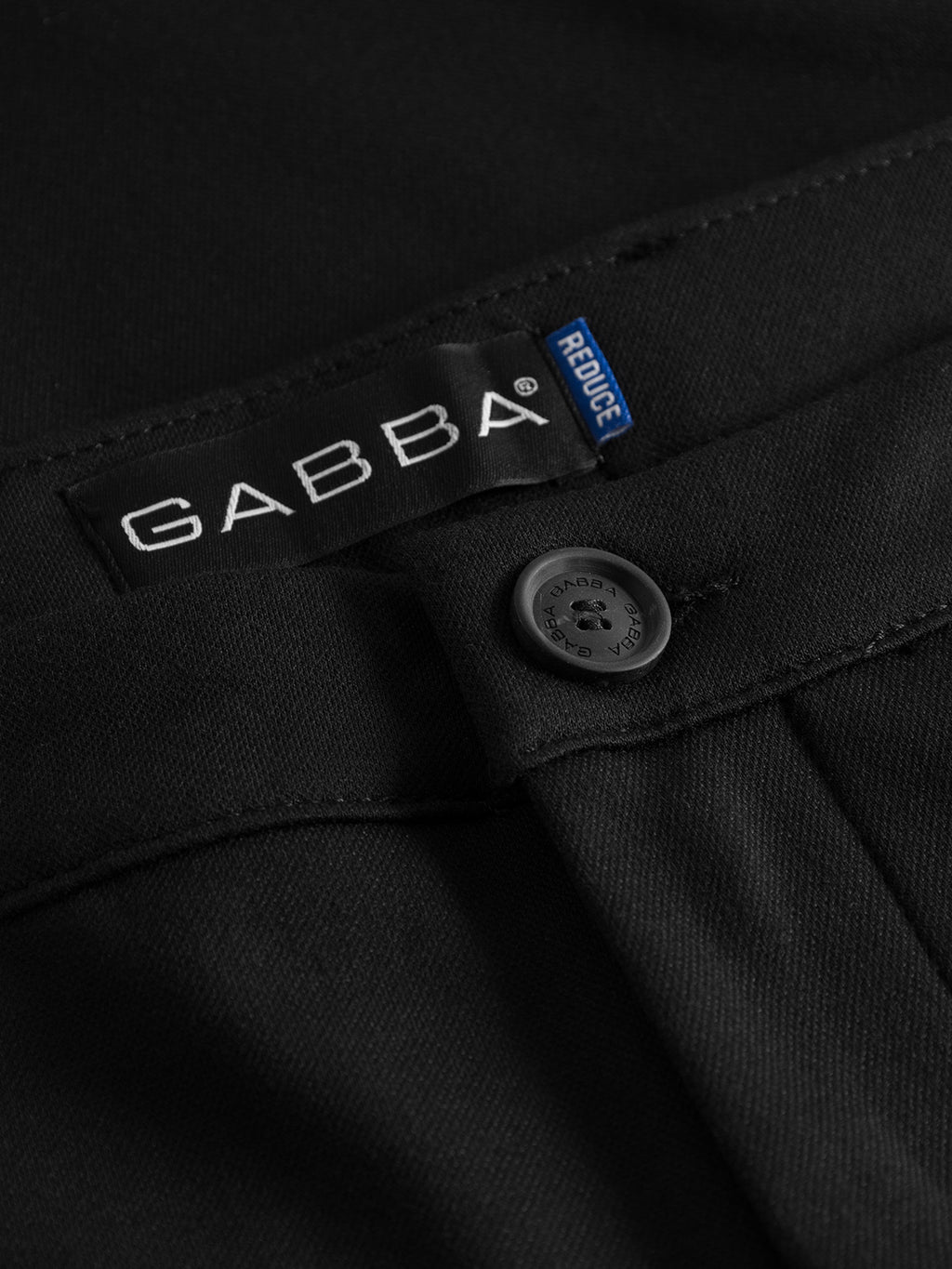 Close-up of Pisa Jersey Pant - Black by GABBA in a tapered fit, featuring a button and belt loop. Includes a black label with white text and a small blue tag. The high-stretch fabric appears smooth and well-tailored, perfect for that metro look.