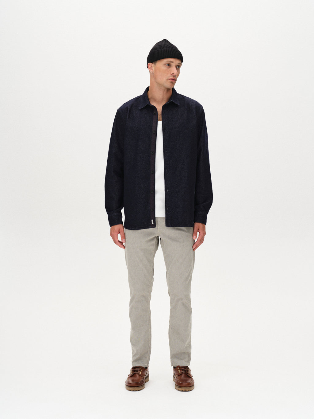 A man wearing a black beanie, dark blue button-up shirt over a white tee, Gabba Paul K3280 Dale Chino in Birch, and brown boat shoes stands against a plain white background, looking slightly to his left.