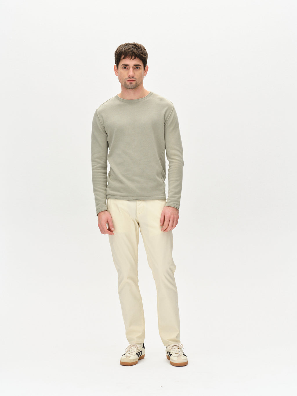 A man stands against a white background, wearing an olive green long-sleeve shirt and Gabbas Paul K3280 Dale Chino in ecru light with French pockets. Beige sneakers complete his look. His hands are by his sides, and he has short, dark hair.