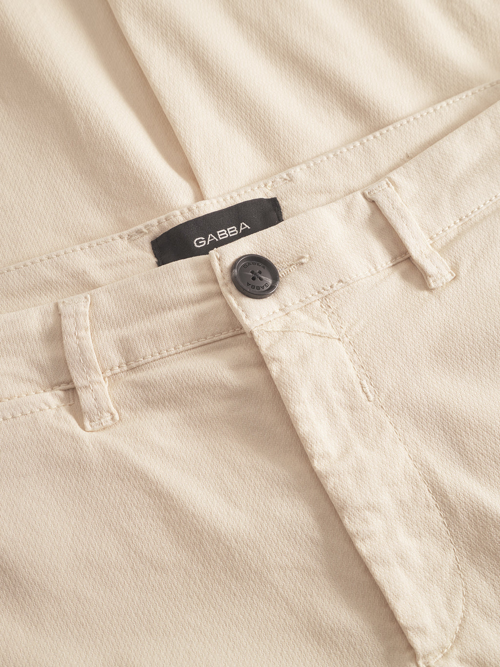 Close-up of the Paul K3280 Dale Chino in Ecru Light by Gabba. These beige slim chinos feature a black branded button, belt loops, and a black Gabba label inside the waistband. They have a soft texture, French pockets, and are neatly folded.