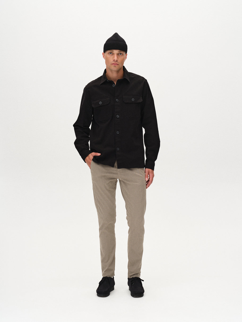 A man poses against a white background in a black beanie, black button-up shirt with branded buttons, GABBA Paul K3280 Dale Chino in Humus, and black shoes. His left hand is in his pocket and he maintains a neutral expression.