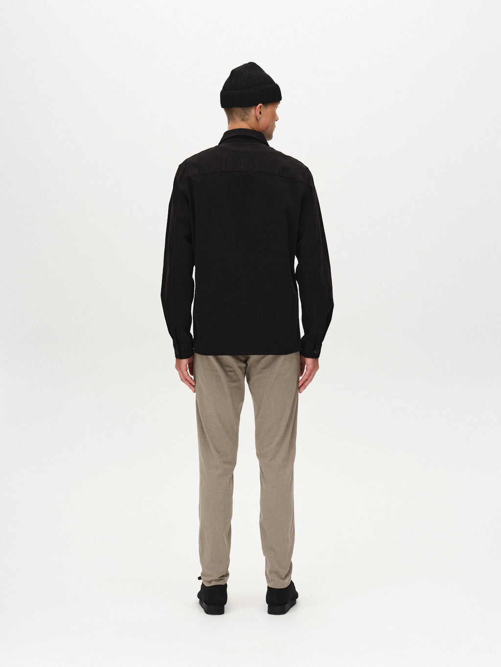 A man stands with his back to the camera, wearing a black jacket, GABBA Paul K3280 Dale Chino - Humus pants with French pockets and branded buttons, black shoes, and a black beanie against a plain white background.