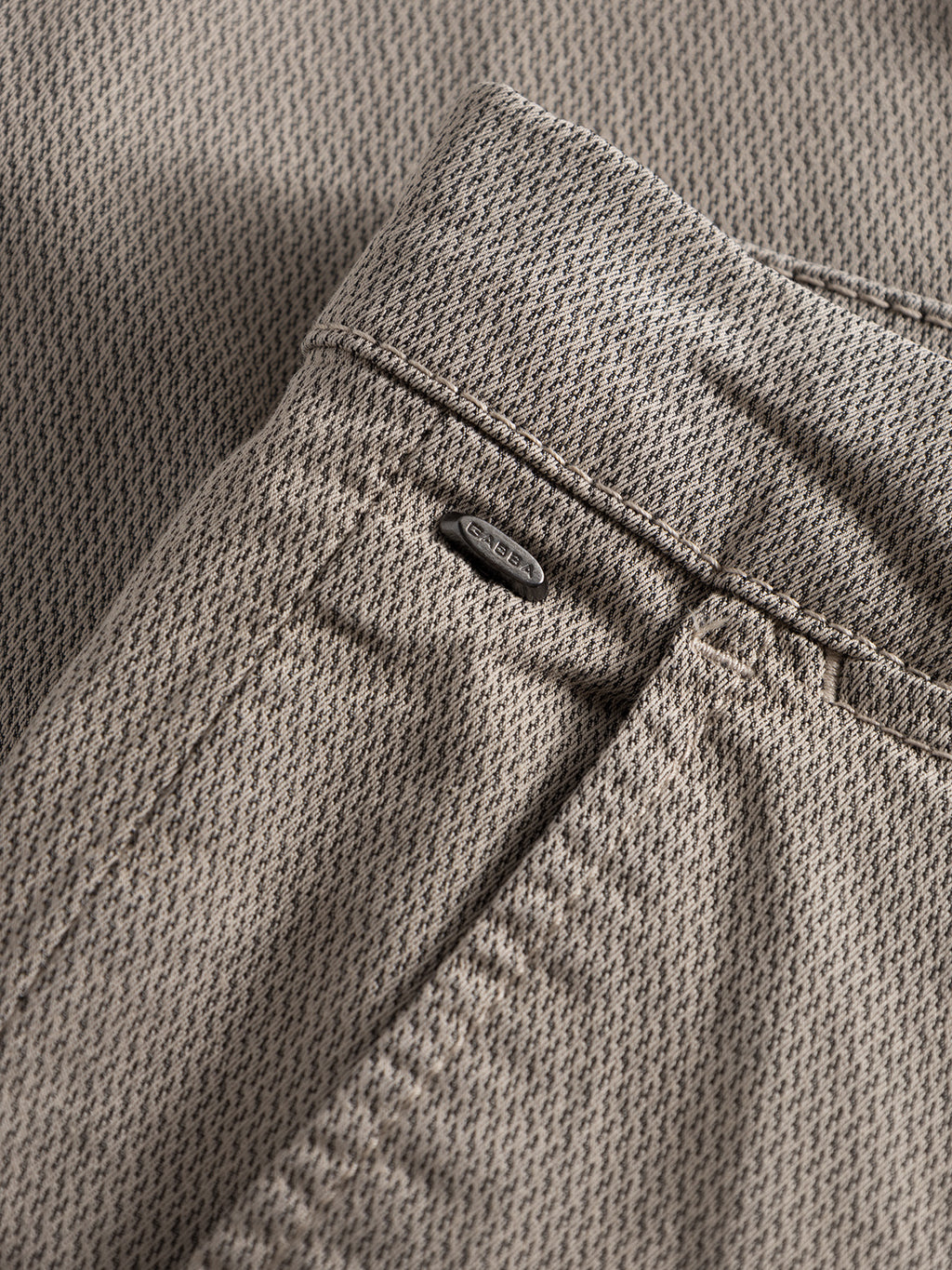 Close-up of the Paul K3280 Dale Chino pants in humus by GABBA, highlighting a small metal brand tag by the French pocket. The fabric features a subtle, repeating pattern.