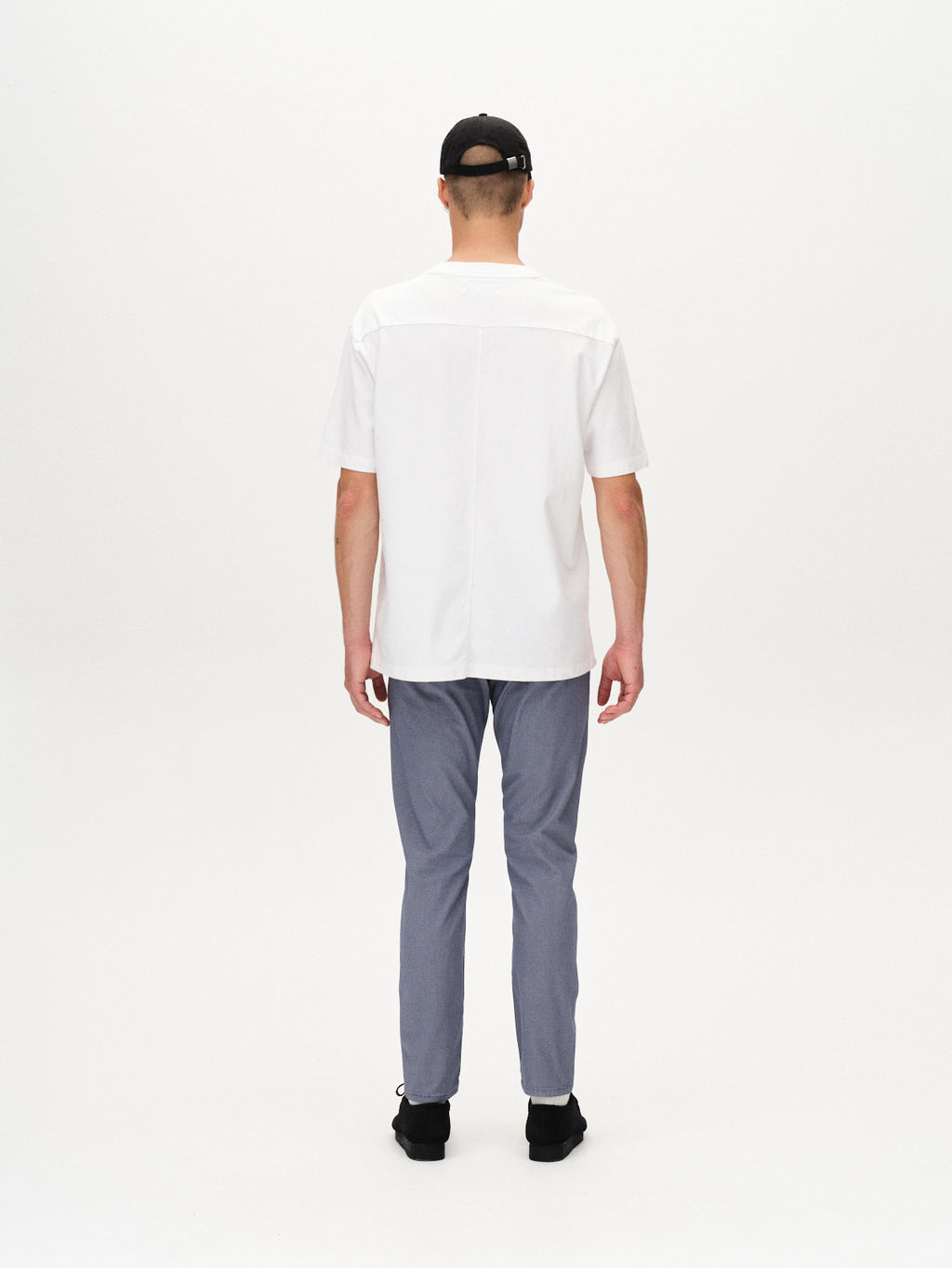 A person stands facing away from the camera, wearing a black baseball cap, a white short-sleeve shirt with branded buttons, Gabba Paul K3280 Dale Chino in blue, and black shoes against a plain white background.