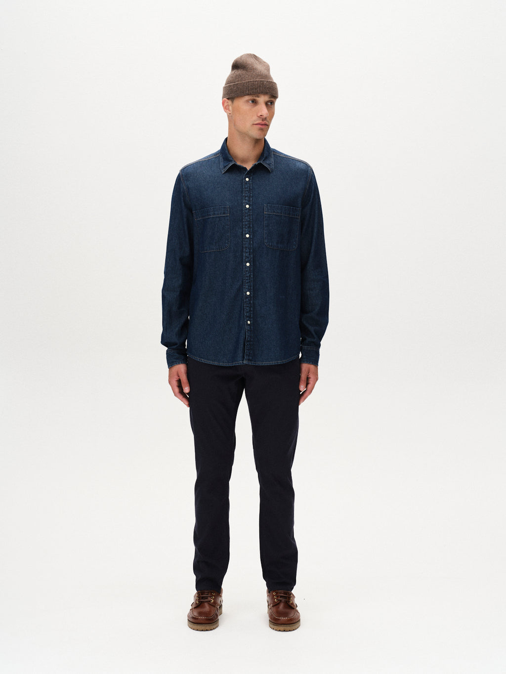 A man stands against a white background, wearing a brown beanie, dark denim button-up shirt, and GABBA Paul K3280 Dale Chino in Navy, showcasing modern style with French pocket slim chinos.