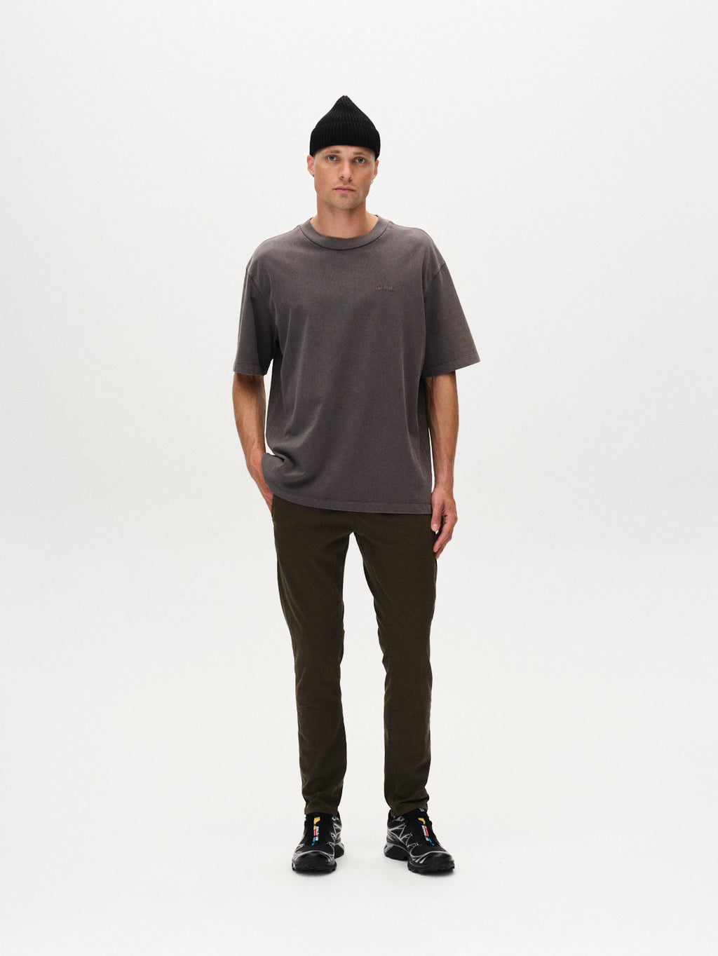 A person stands facing forward against a white background, wearing a black beanie, loose dark gray T-shirt, black sneakers, and GABBA’s Paul K3280 Dale Chino in Army. One hand is in their pocket and they have a neutral expression.