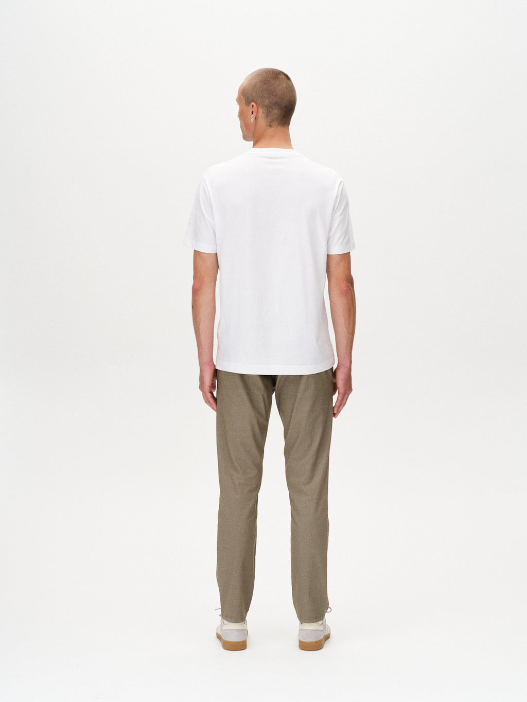 A man faces away from the camera, wearing a white short-sleeve t-shirt and Gabba Paul K3280 Dale Chino in olive green with French pockets and branded buttons, paired with white sneakers, against a plain white background.