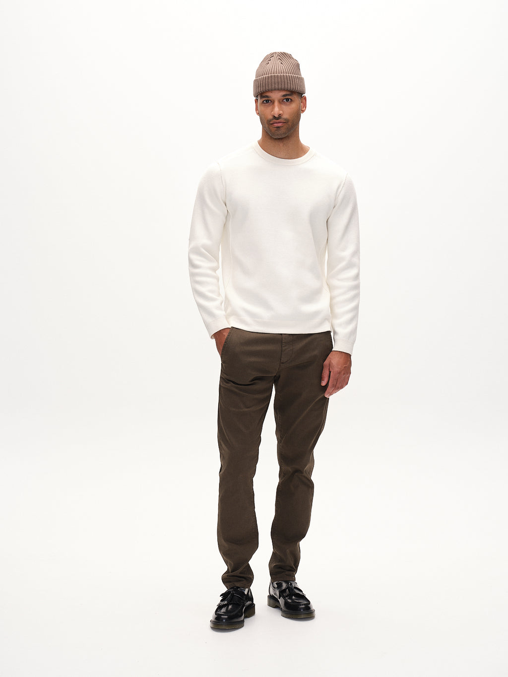 A person in a white long-sleeve shirt, DK. Brown Paul K3280 Dale Chino by Gabba with French pockets, and black shoes stands against a plain white background. They wear a beige knit beanie and have a neutral expression, embodying Gabba’s effortless elegance.