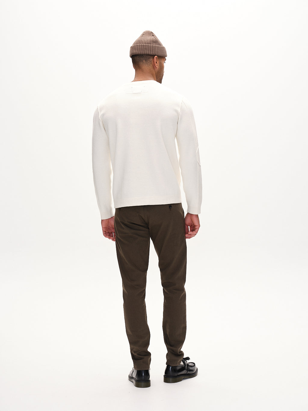 A person stands facing away, wearing a beige knit beanie, a white sweater, and Gabbas Paul K3280 Dale Chino in dark brown. Black shoes complete the look against a plain white background, enhancing the minimalist style.