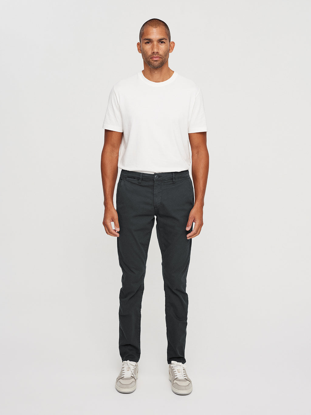 Facing forward against a plain background, a person wears a white T-shirt paired with GABBAs Paul K3280 Dale Chino in grey. Light-colored sneakers enhance the look, while branded buttons add detail. Their arms rest naturally by their sides as they maintain a neutral expression.