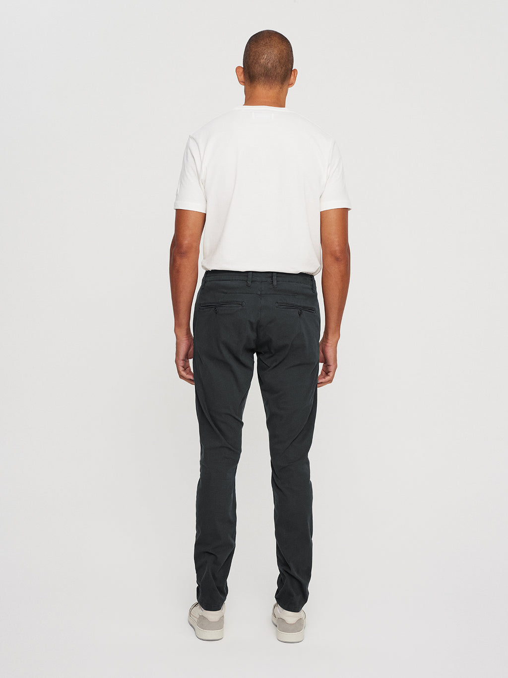 A person stands against a white backdrop wearing a white T-shirt, GABBAs Paul K3280 Dale Chino in grey with French pockets, and light shoes. Their arms rest by their sides.