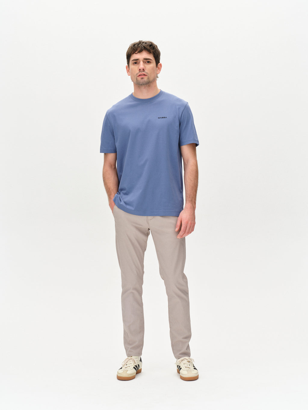 A man stands facing forward in a plain studio, wearing a blue t-shirt, Gabba Paul K3280 Dale Chino in Grey Paloma with French pockets, and cream sneakers with black accents. He has a neutral expression and relaxed arms at his sides.