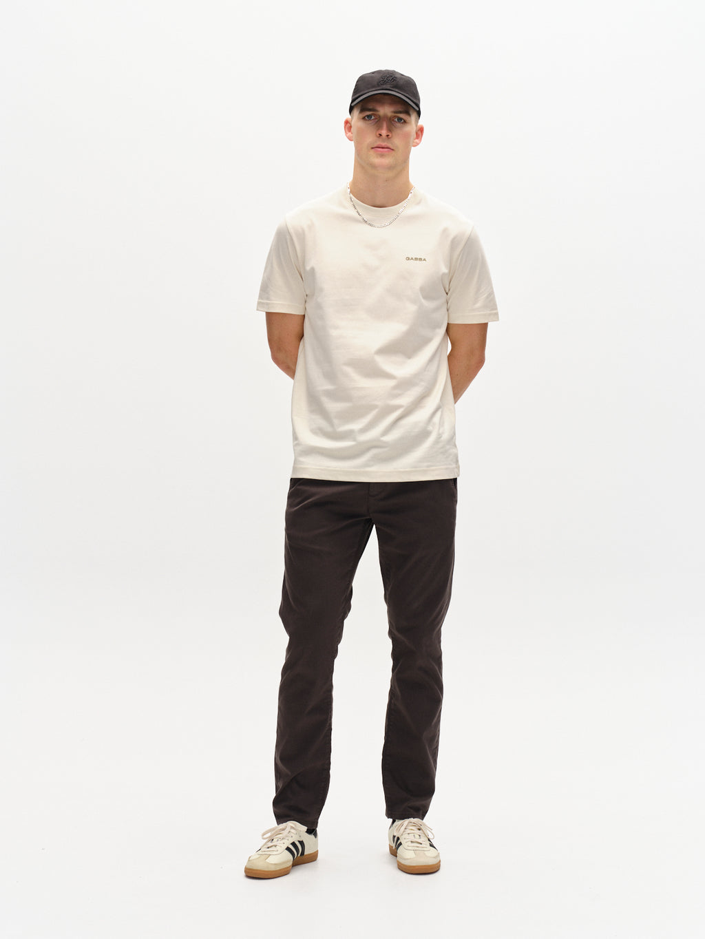 A person poses against a plain white background, wearing a black cap, white t-shirt, Paul K3280 Dale Chino - Charcoal with French pockets, and white sneakers with black stripes. Their hands are behind their back, exuding a casual style inspired by Gabba.