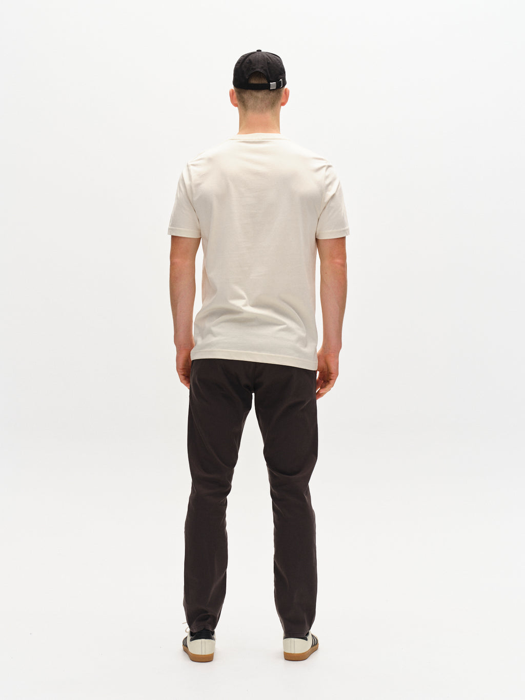 A person faces away from the camera in a beige T-shirt, wearing Gabbas Paul K3280 Dale Chino pants in charcoal with French pockets, a black cap, and sneakers against a plain white backdrop.