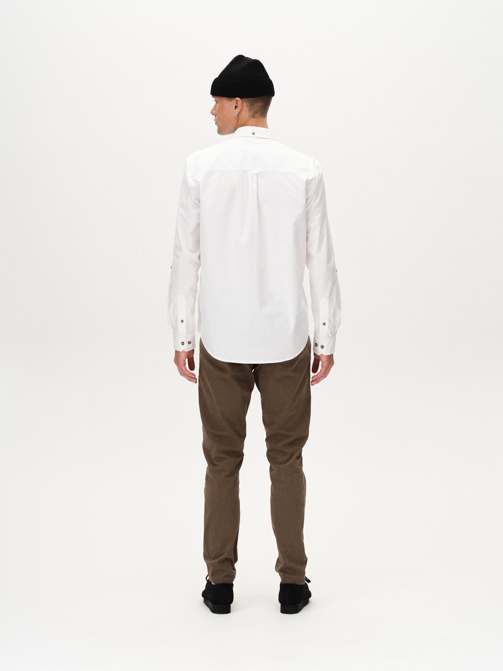 A man wearing a black beanie, white long-sleeve button-up, Gabba Paul K3280 Dale Chino - Crocodile pants with French pockets, and black shoes stands facing away against a plain white background.