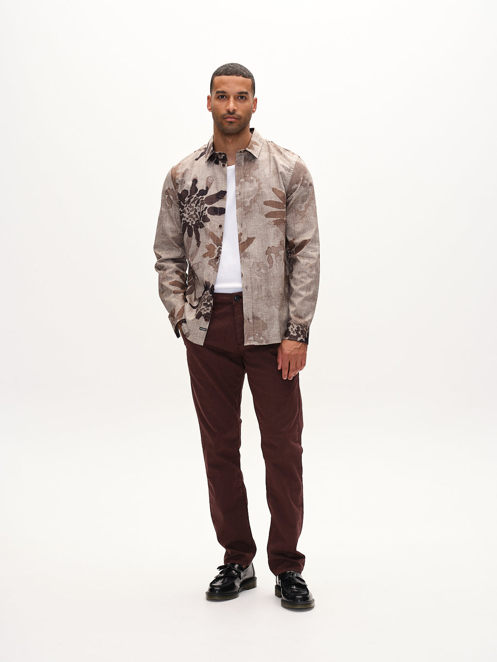 A man stands against a plain background wearing a beige patterned shirt over a white tee, with hands in the French pockets of his Gabba Paul K3280 Dale Chino pants in Burnt Purple. He’s wearing black shoes with branded buttons and looking at the camera.