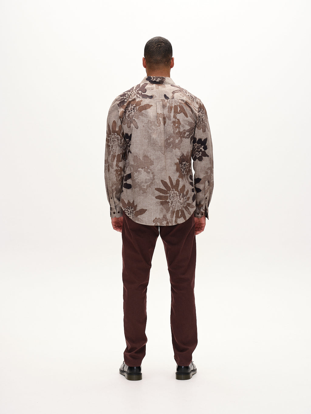 A person stands facing away, wearing a floral-patterned shirt in shades of brown, beige, and black. They pair it with Gabbas Paul K3280 Dale Chino in Burnt Purple, featuring French pockets and branded buttons. Black shoes complete the look against a plain white background.