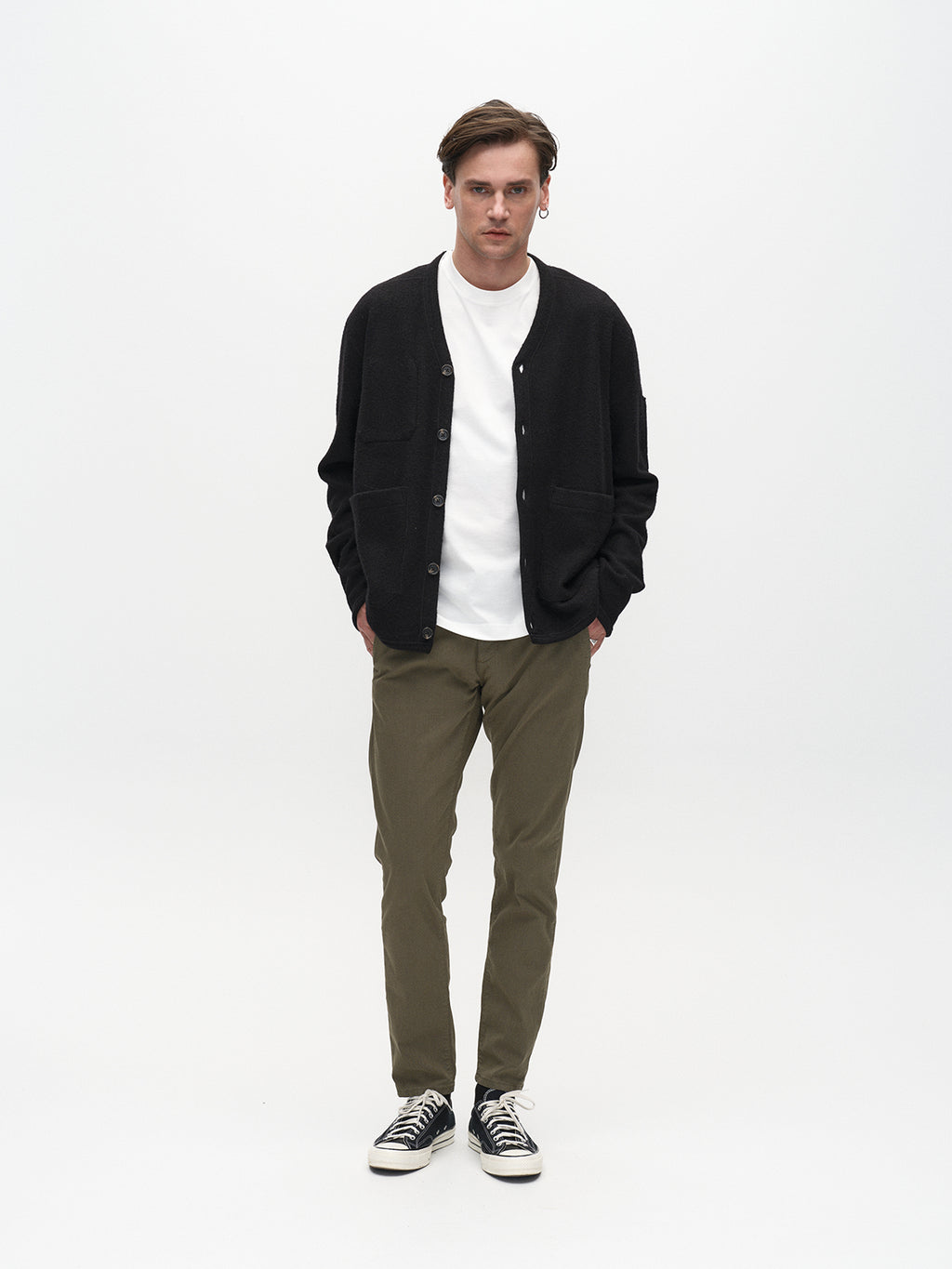 Against a white backdrop, a person epitomizes classic style in a black cardigan and a white shirt. They wear Gabbas Paul K3280 Dale Chino in Deep Lichen Green with French pockets, paired with black sneakers. With short hair and forward gaze, their hands rest casually in their pockets.