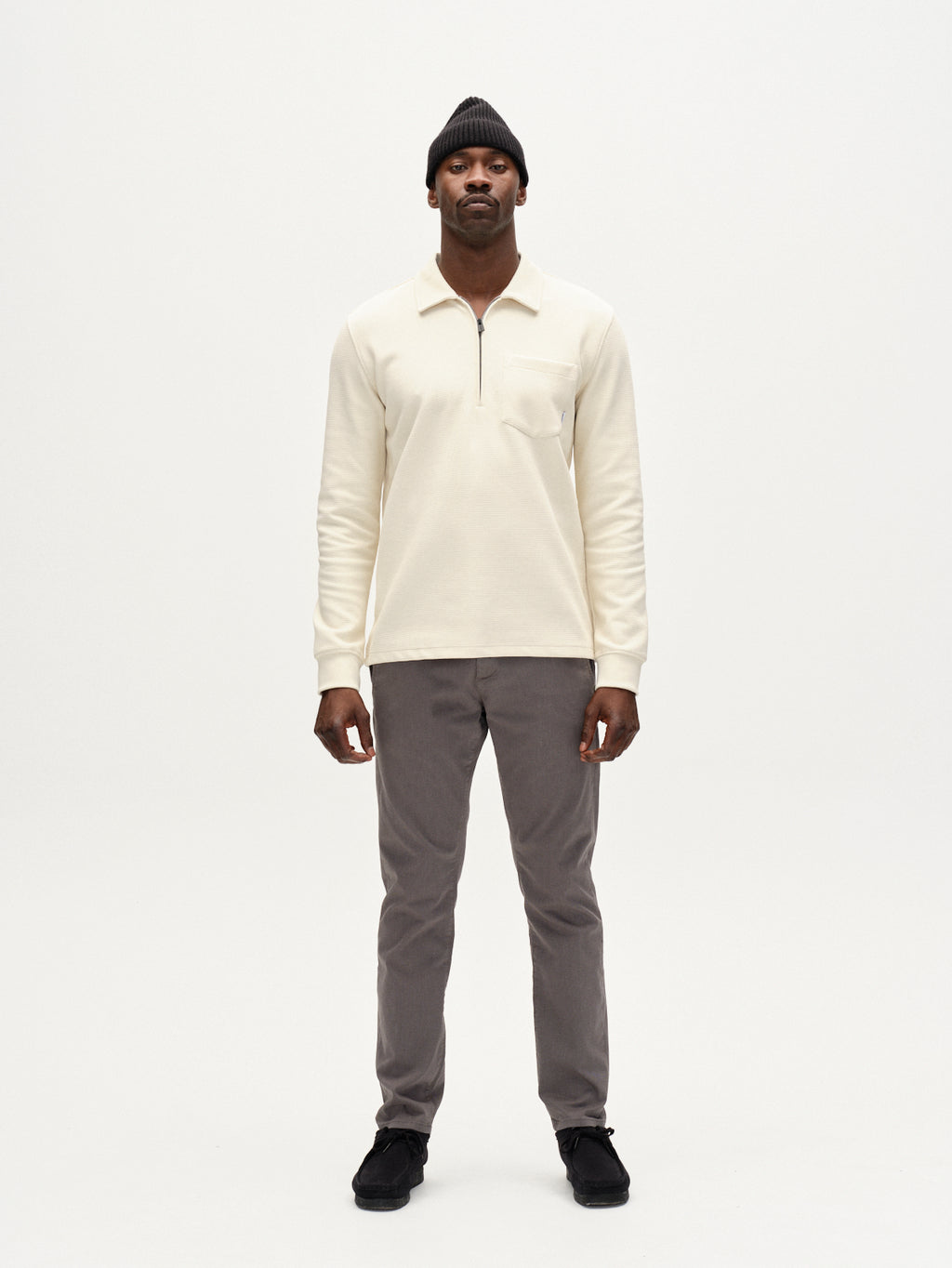 A man stands facing forward against a white background, wearing a cream long-sleeve zip-collar shirt, Gabba Paul K3280 Dale Chino in Dust Sage Green, black shoes, and a black knit beanie.